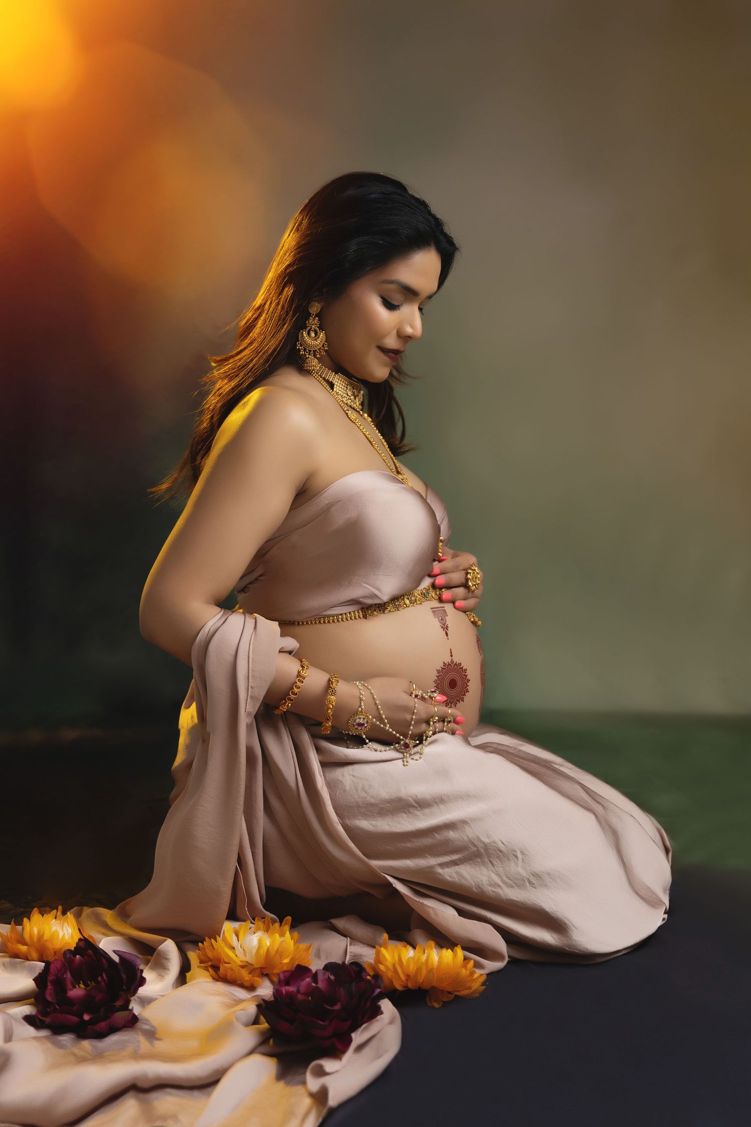 Pregnant woman in blush sari with gold jewelry kneels gracefully among flowers in a warm, glowing maternity portrait.