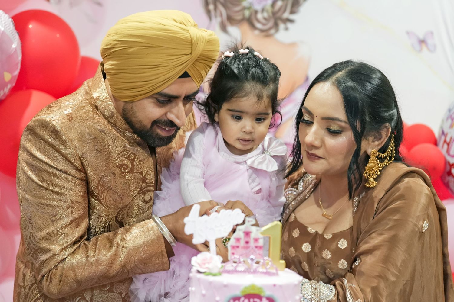 South Asian family celebrating baby girl's first birthday with pink cake, parents in traditional attire.