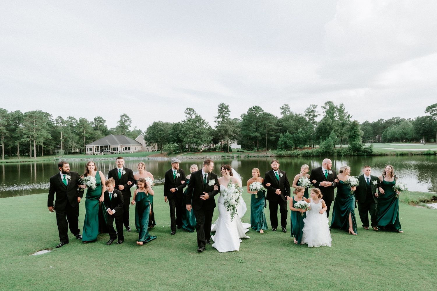 Wedding party in emerald green and black attire poses on a golf course with a lake and trees in the background.
