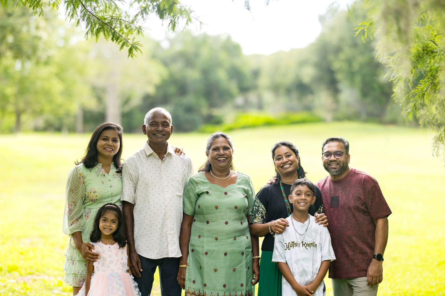 Multi-generational Indian family smiling together outdoors in a lush green park on a sunny day.
