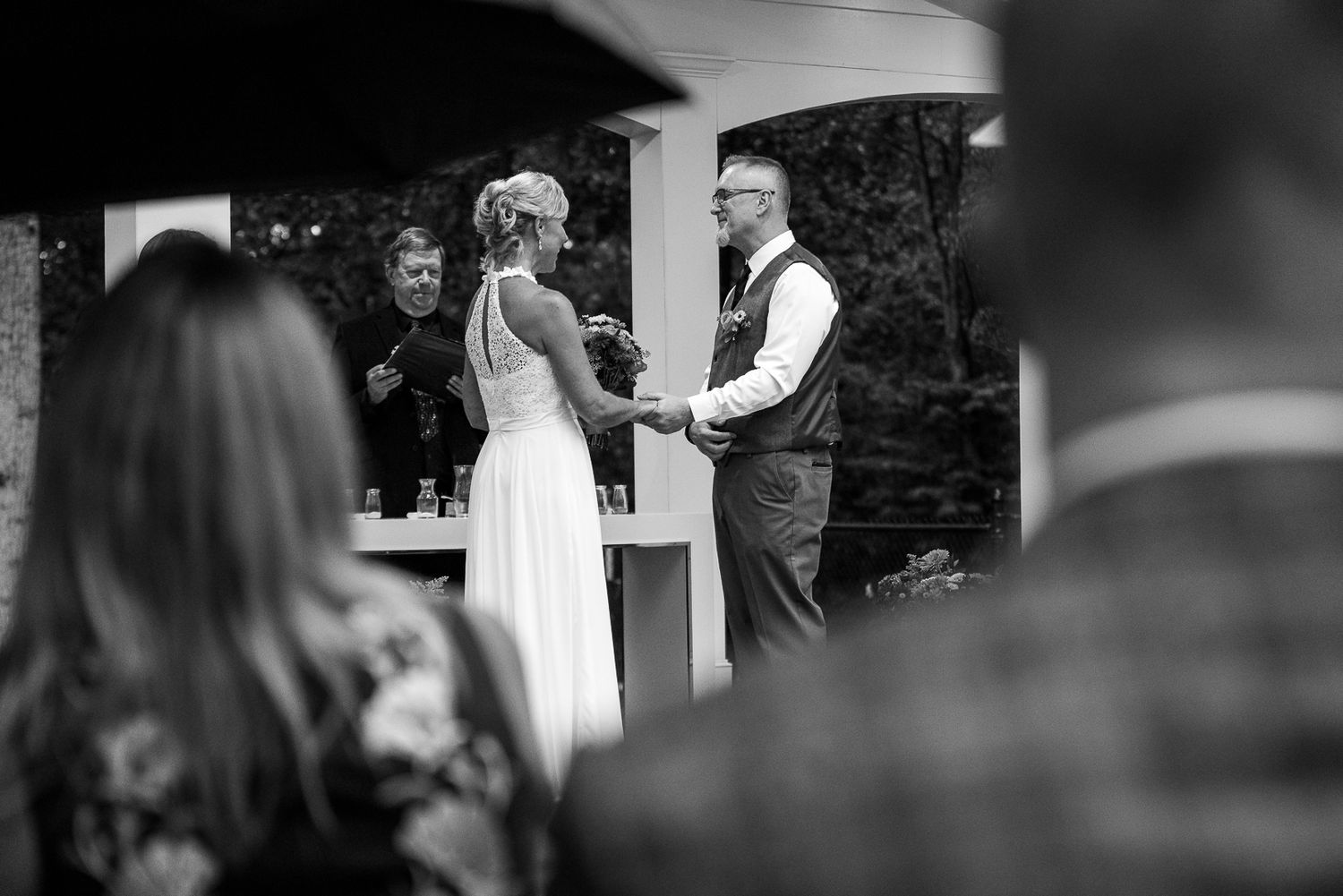 Black and white wedding ceremony photo of bride and groom exchanging vows outdoors with officiant.