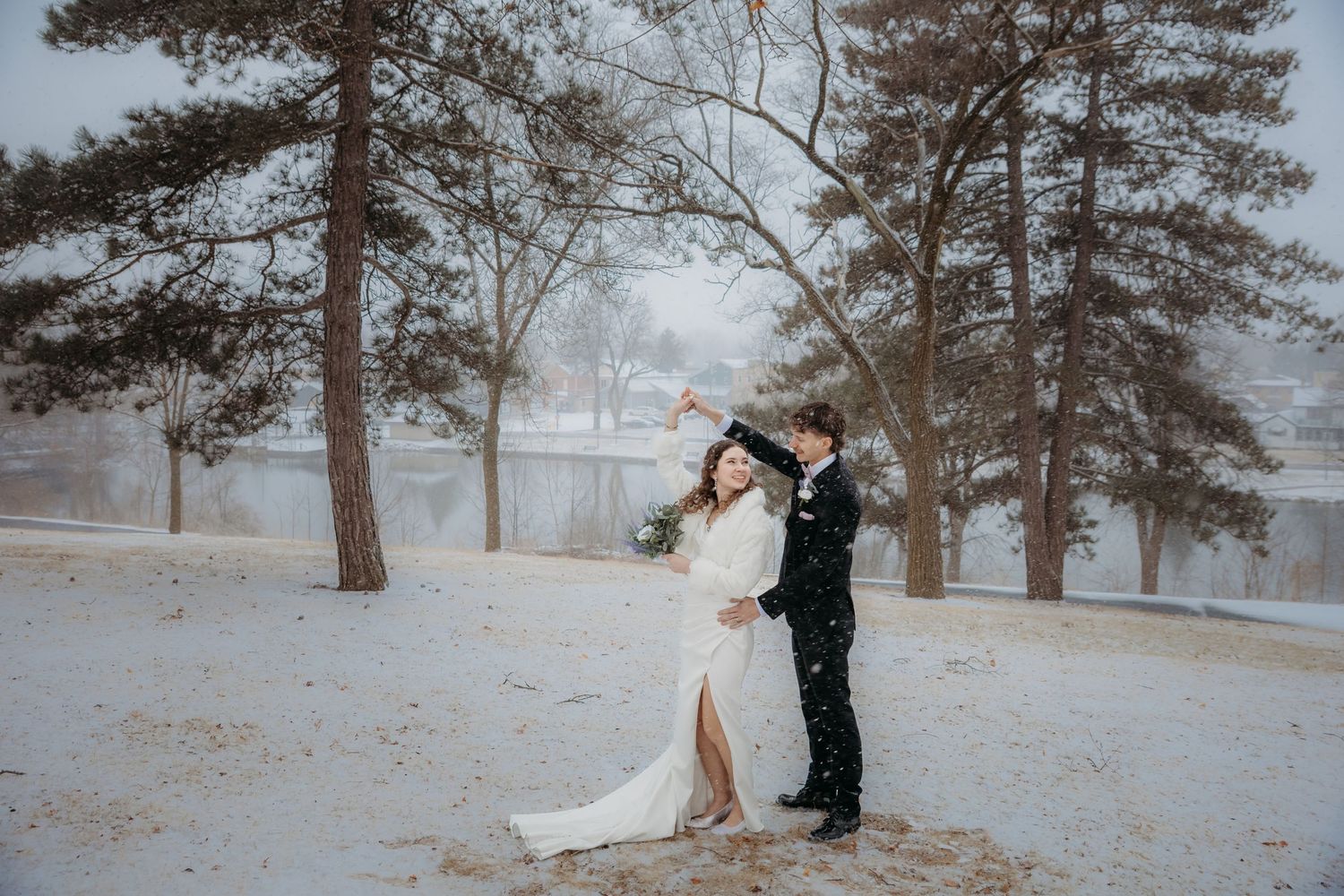 Bride and groom dancing in snowy winter landscape with snow-covered trees and frozen lake backdrop.