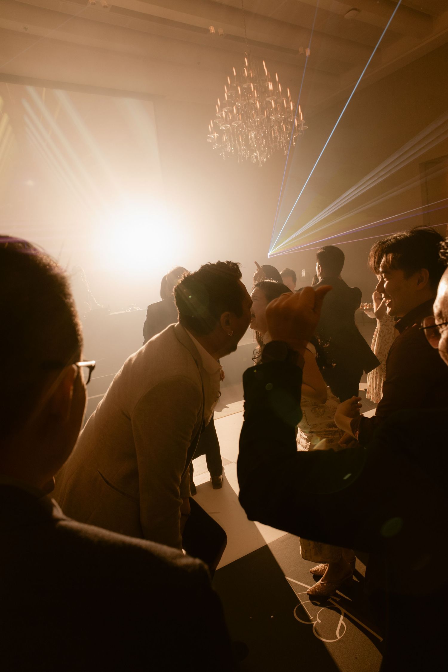 Guests dancing at an elegant party with laser lights and a crystal chandelier in a warmly lit venue.