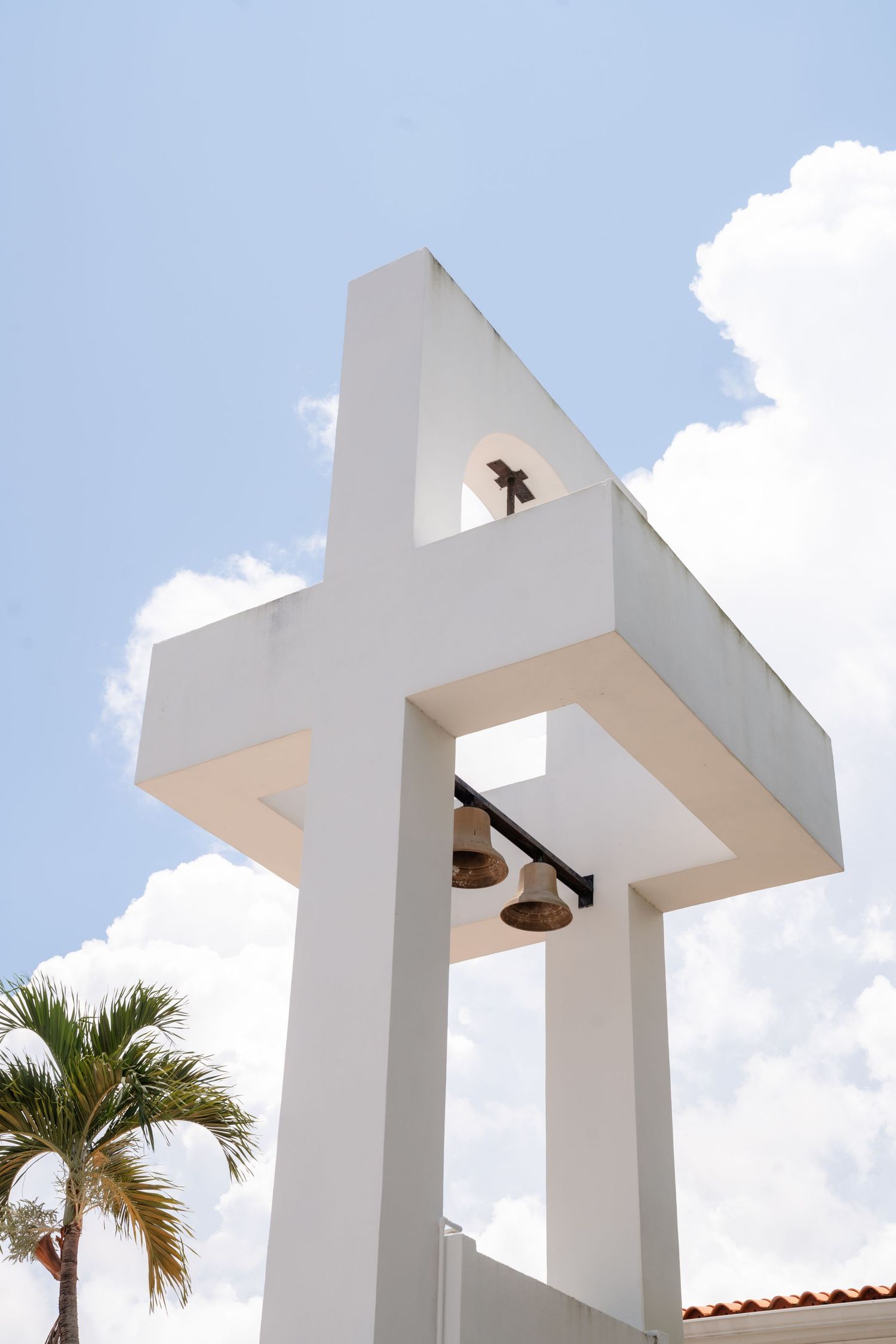 White modern church bell tower with two bronze bells against a blue sky, flanked by a tropical palm tree.