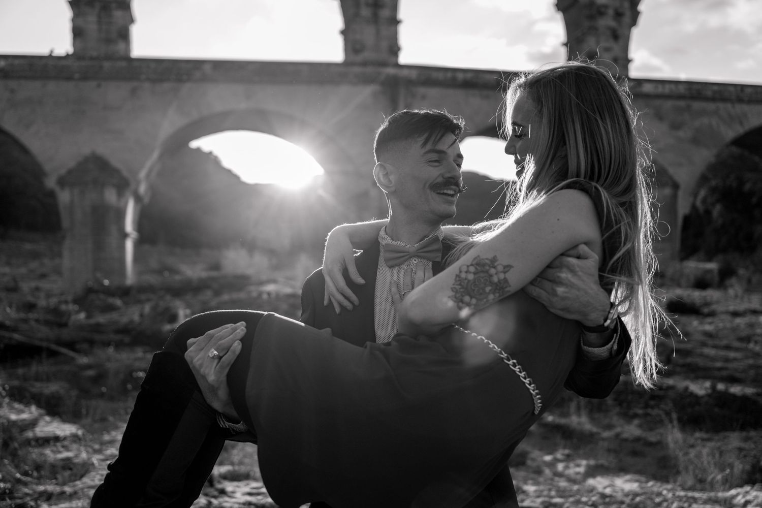 A couple embraces and kisses in silhouette against a dramatic sunset sky with mountains in the background.