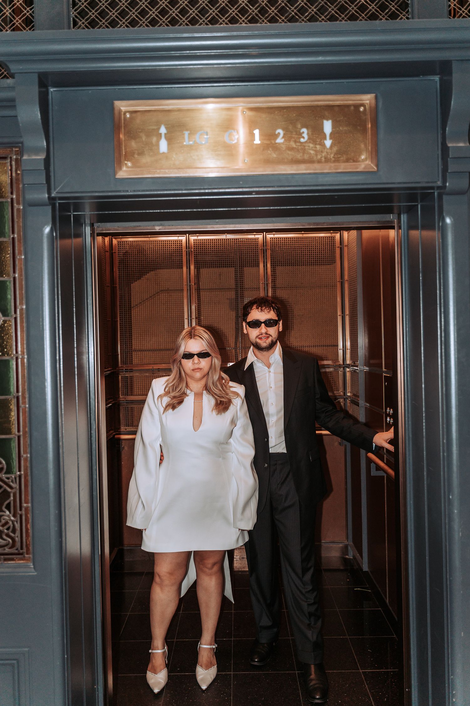Couple in sunglasses posing in an elevator, woman in white mini dress, man in black suit, elegant hotel interior.