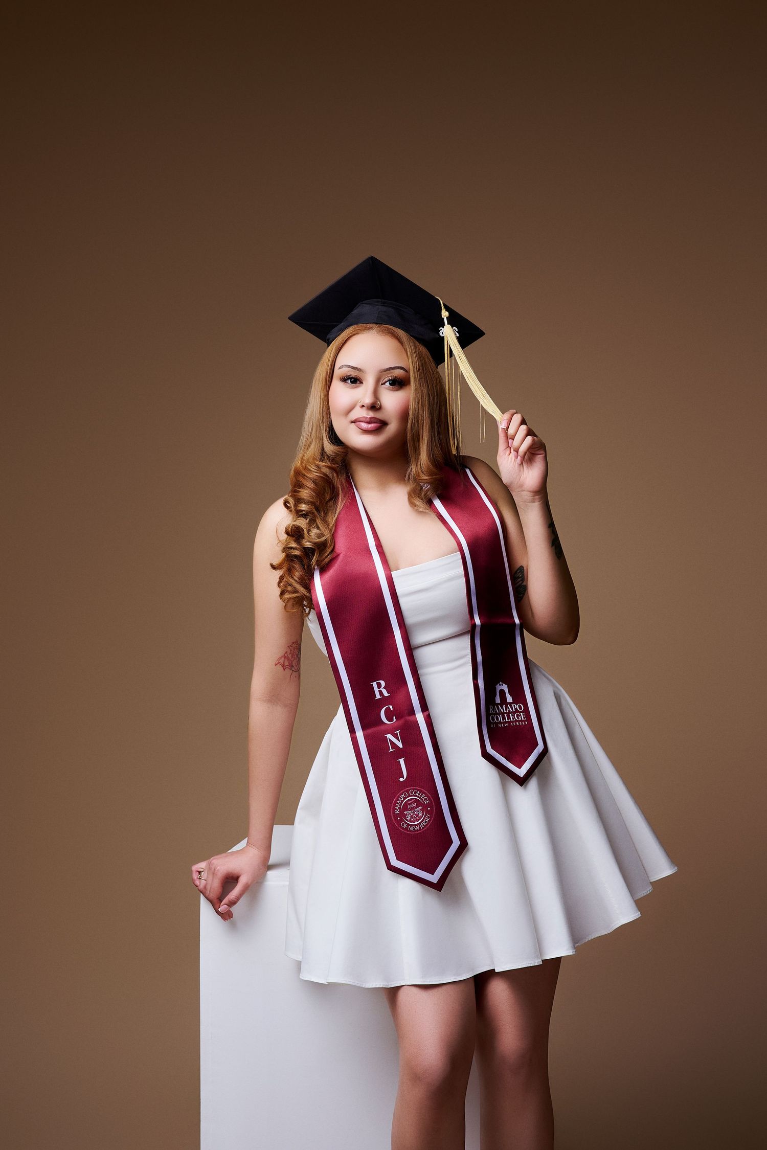 Young woman in white dress and graduation cap wearing a maroon RCNJ sash poses for grad photo.
