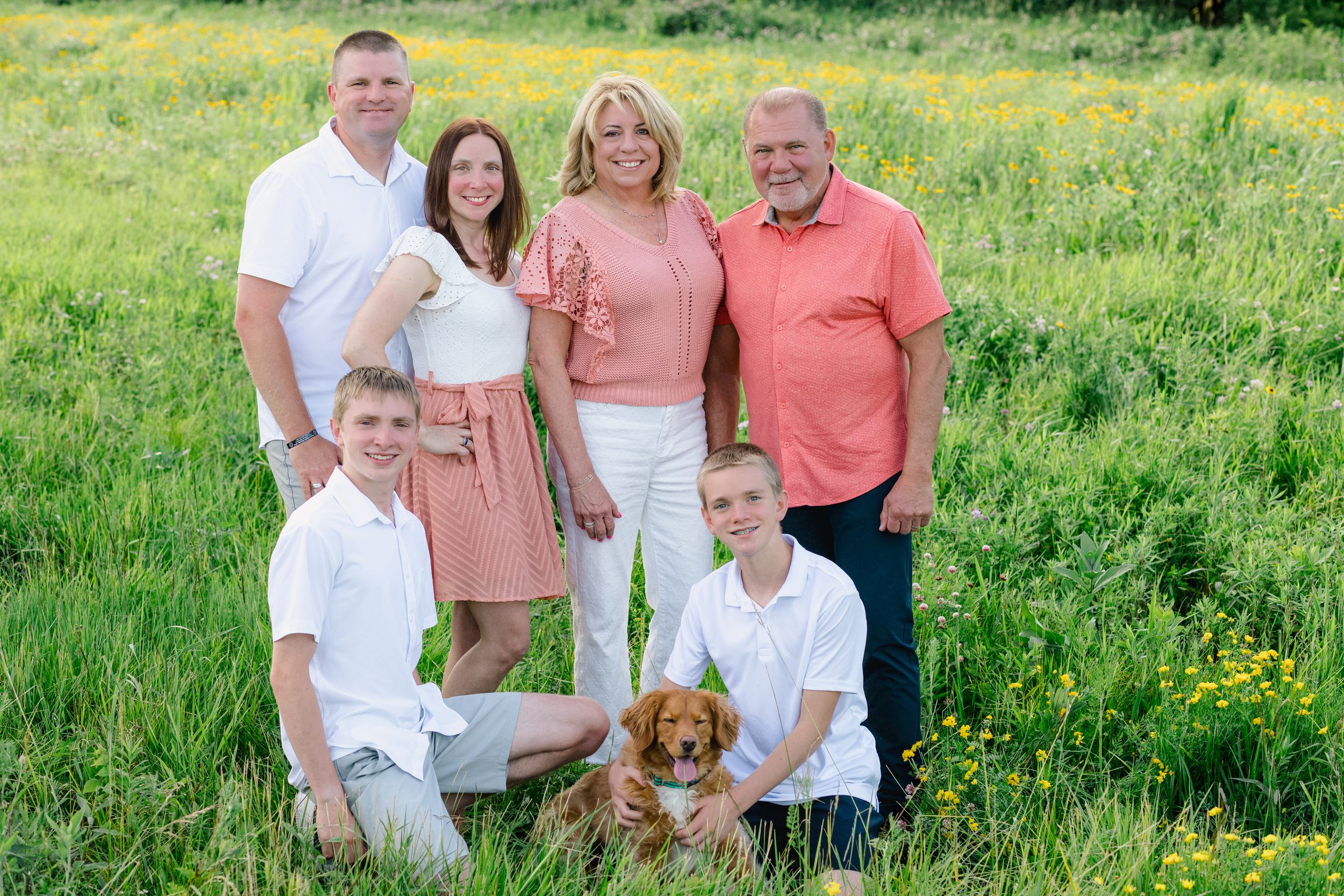 Family of six poses with golden retriever puppy in sunny green meadow with yellow wildflowers.