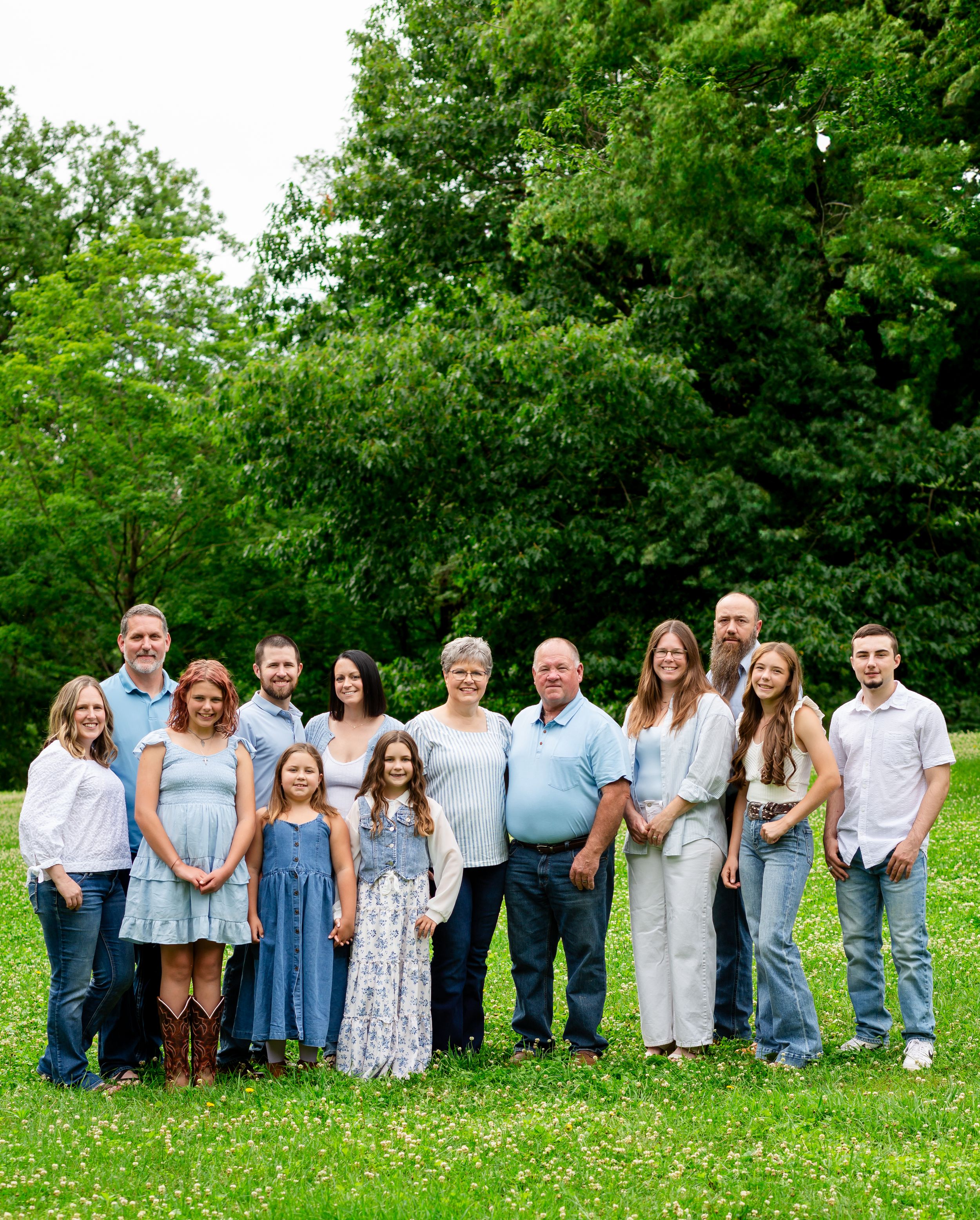 Large family group portrait outdoors on green grass with lush trees, wearing coordinated blue and white outfits.
