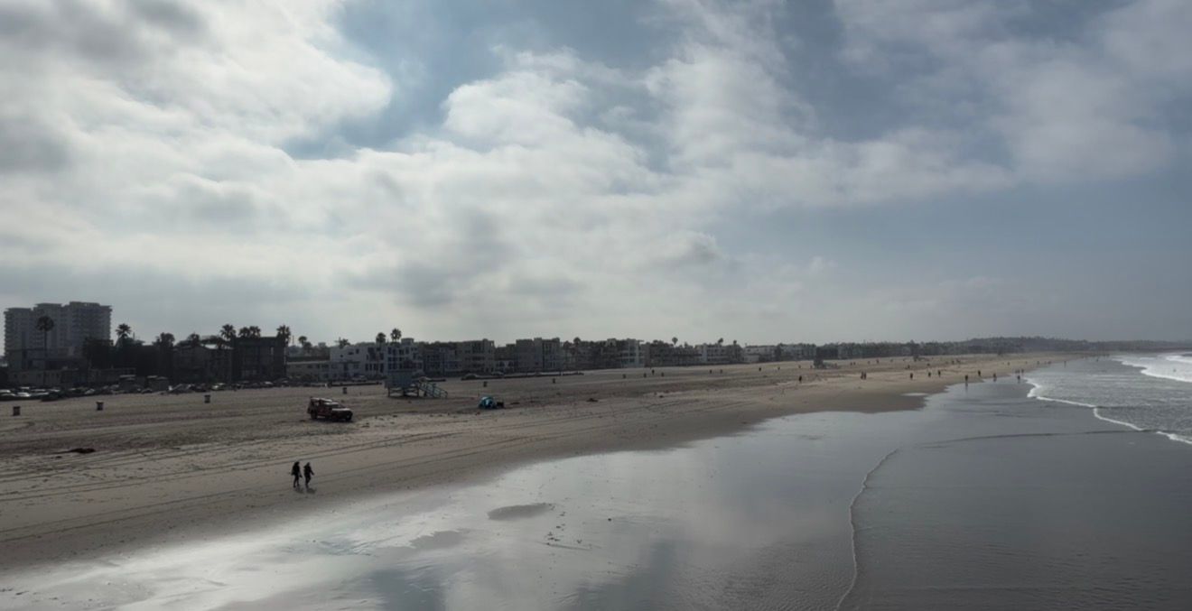 Aerial view of a wide sandy beach with calm ocean waves, cloudy sky, and distant city skyline on a peaceful day.