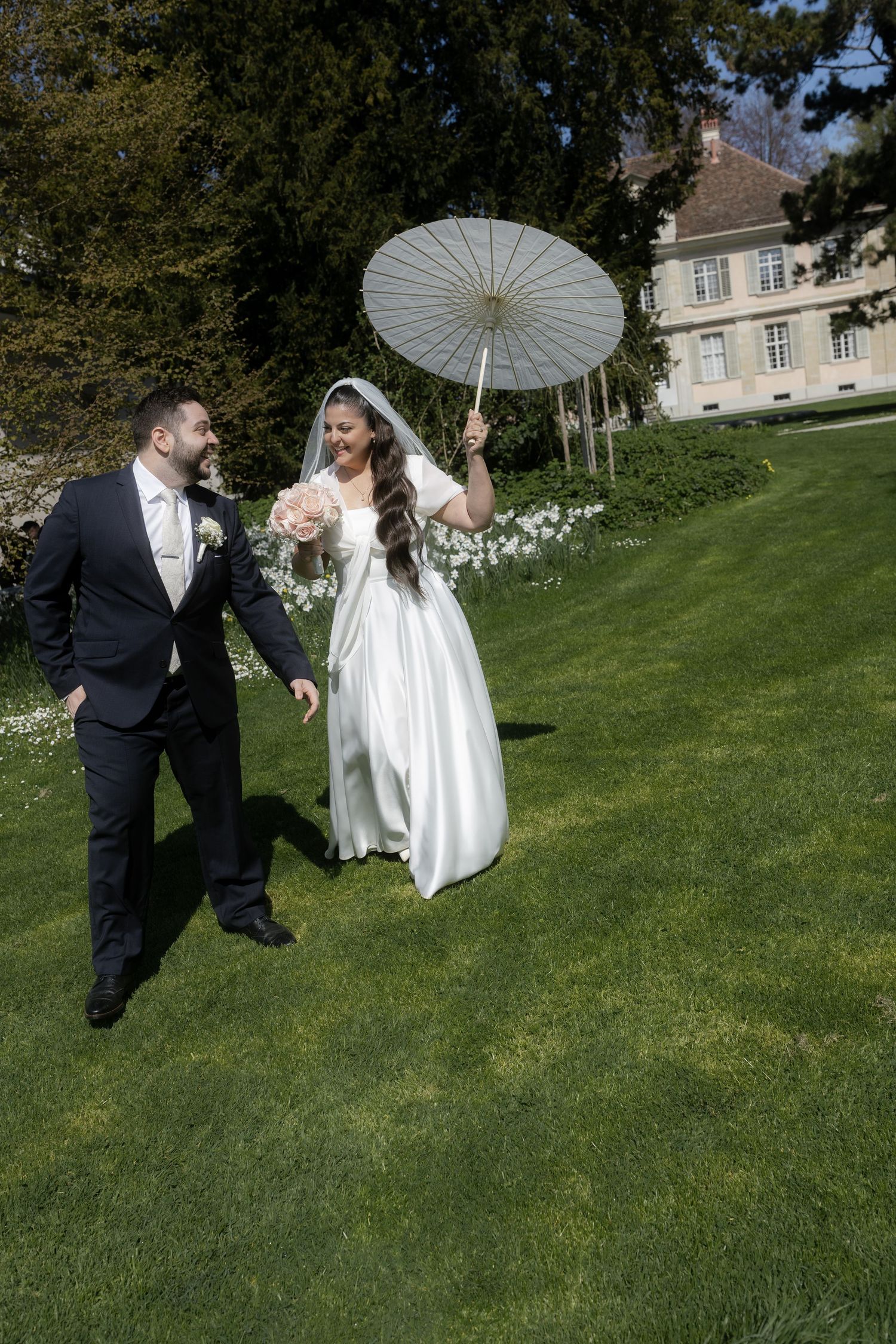Bride in white gown holding parasol walks with groom in black suit on lush green lawn outside elegant estate.
