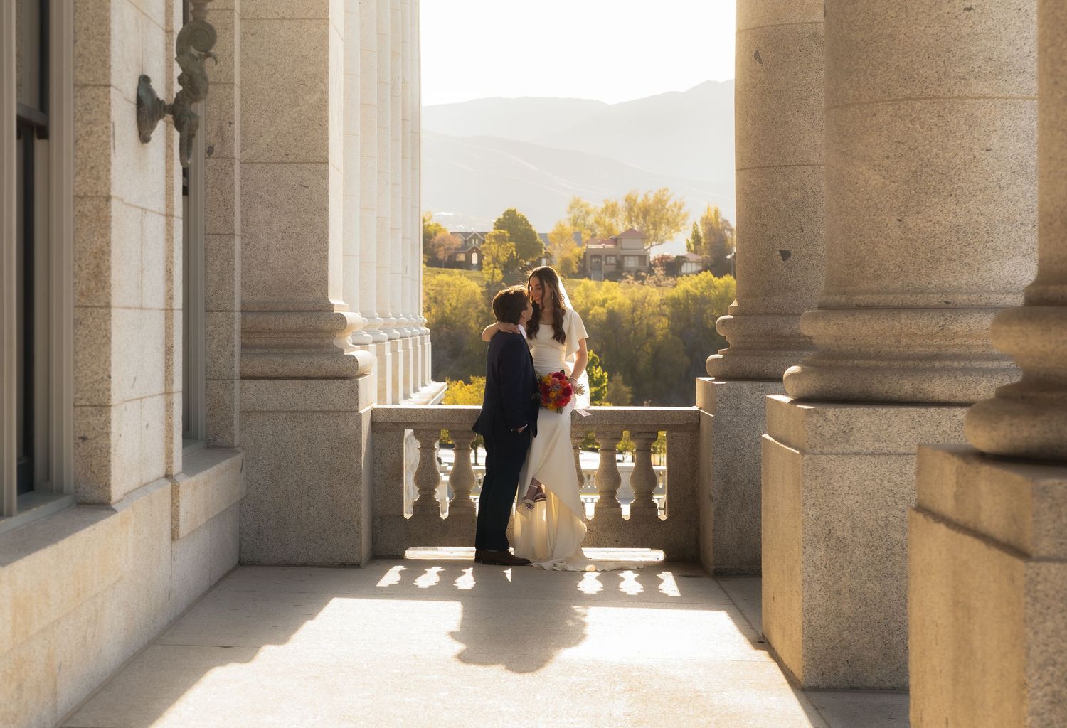 A couple shares a romantic kiss between grand stone columns at a scenic wedding venue with mountain views.
