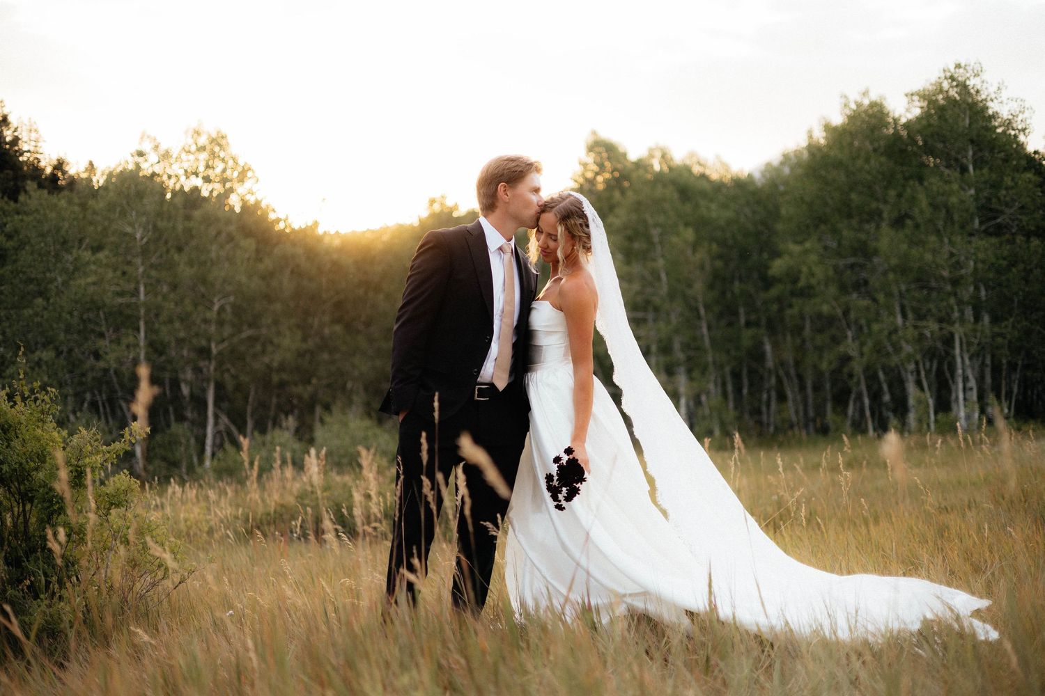 Bride in lace veil holding white flower bouquet at golden sunset in a field.