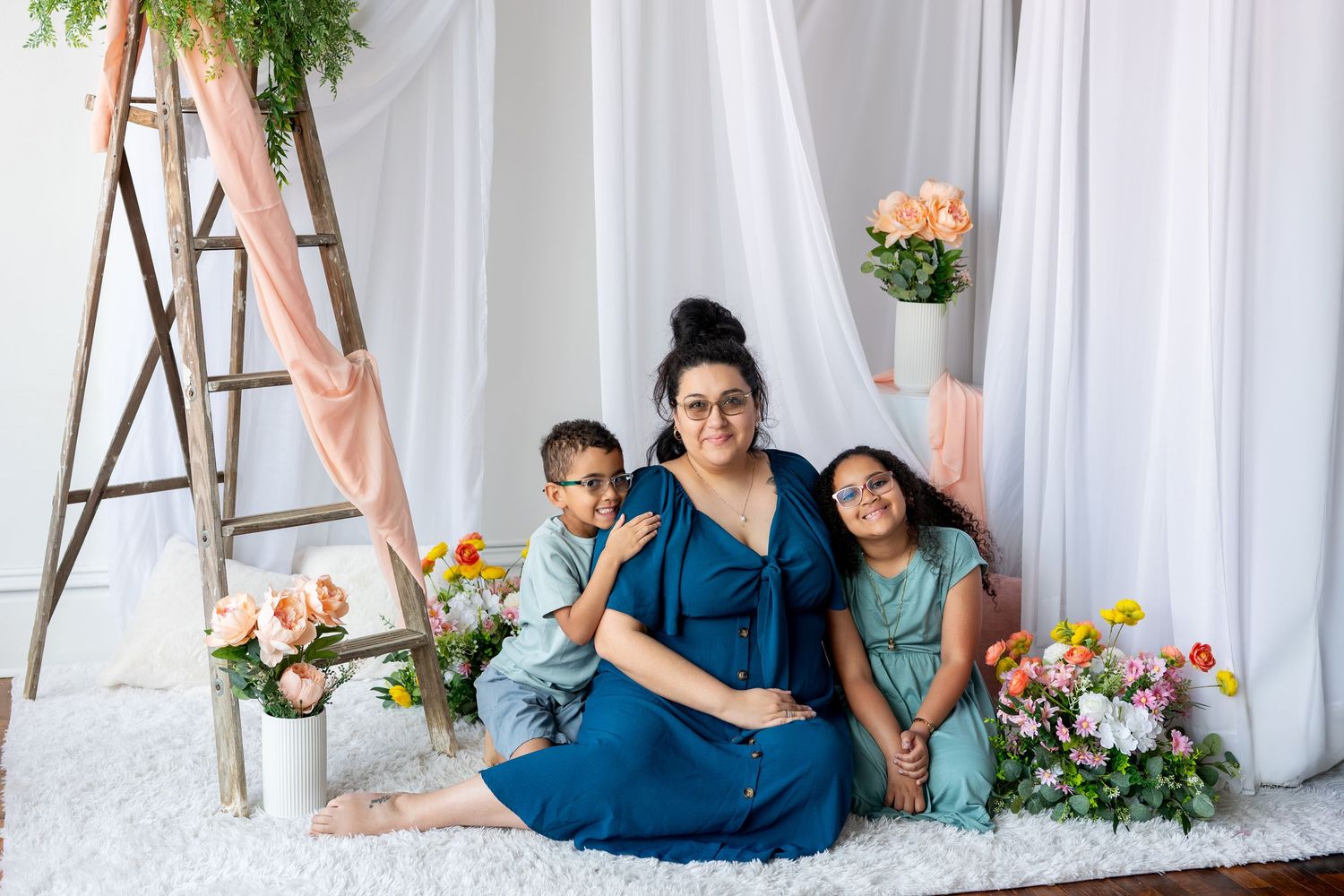 Mother in blue dress sitting with two children surrounded by flowers and white drapes indoors.