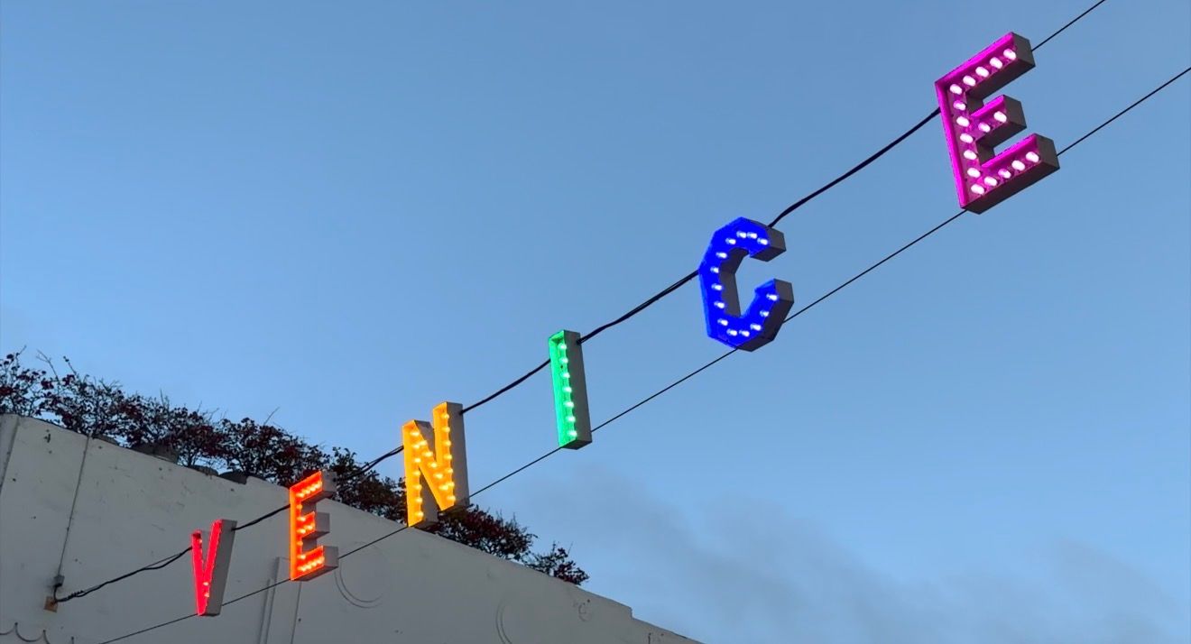 Colorful illuminated VENICE sign letters hanging on wires against a blue sky at dusk in Venice Beach, CA.