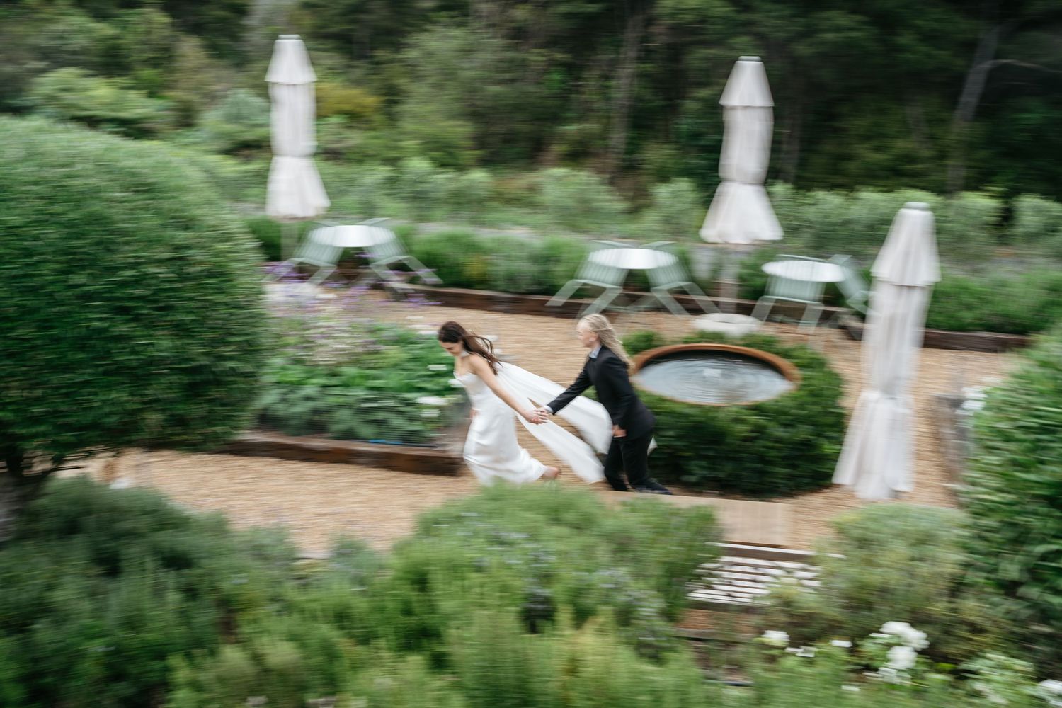 Bride and groom running through lush garden wedding venue with fountain and white patio umbrellas.