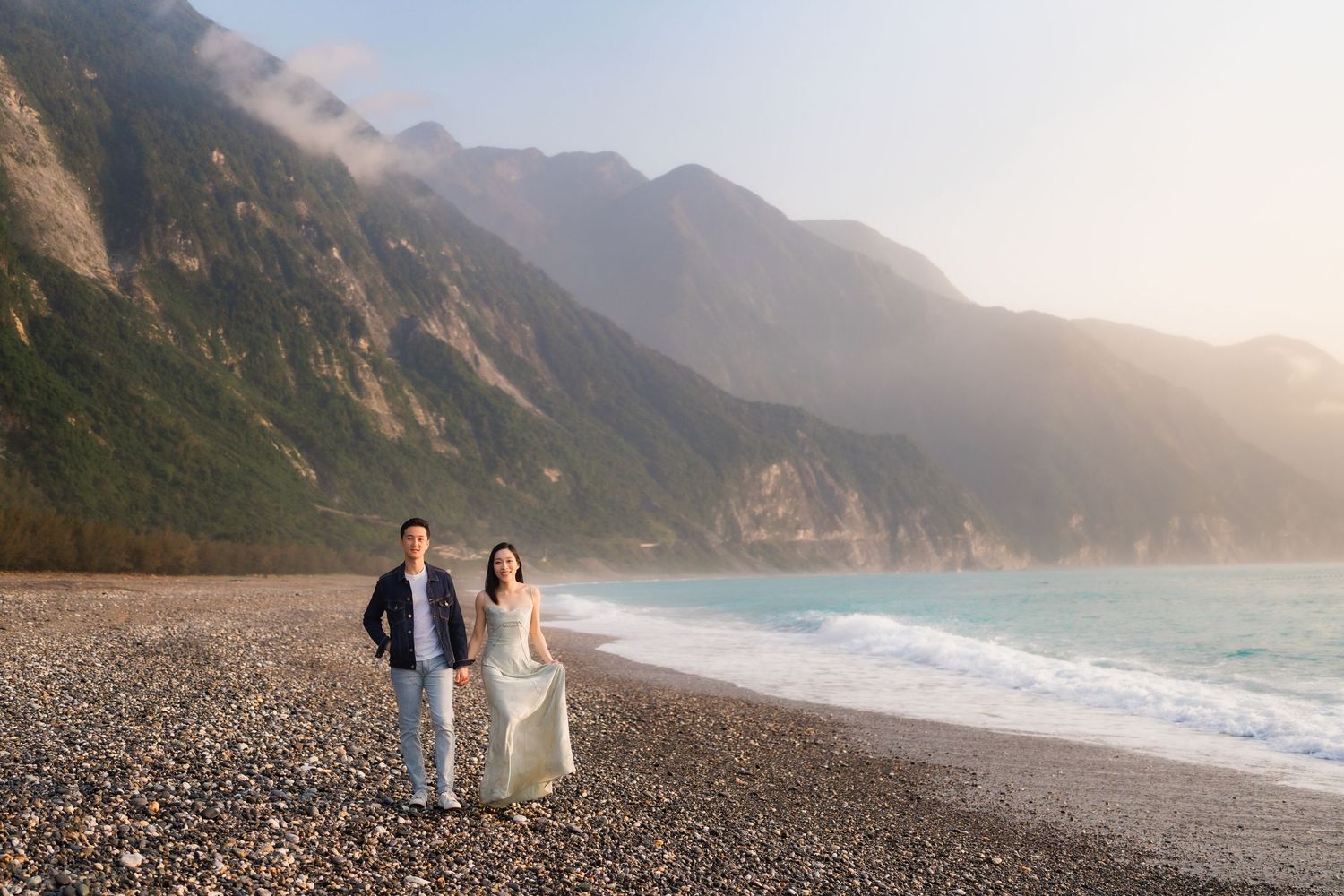 Couple walking hand in hand on a rocky beach with turquoise water and misty green mountains at sunset.