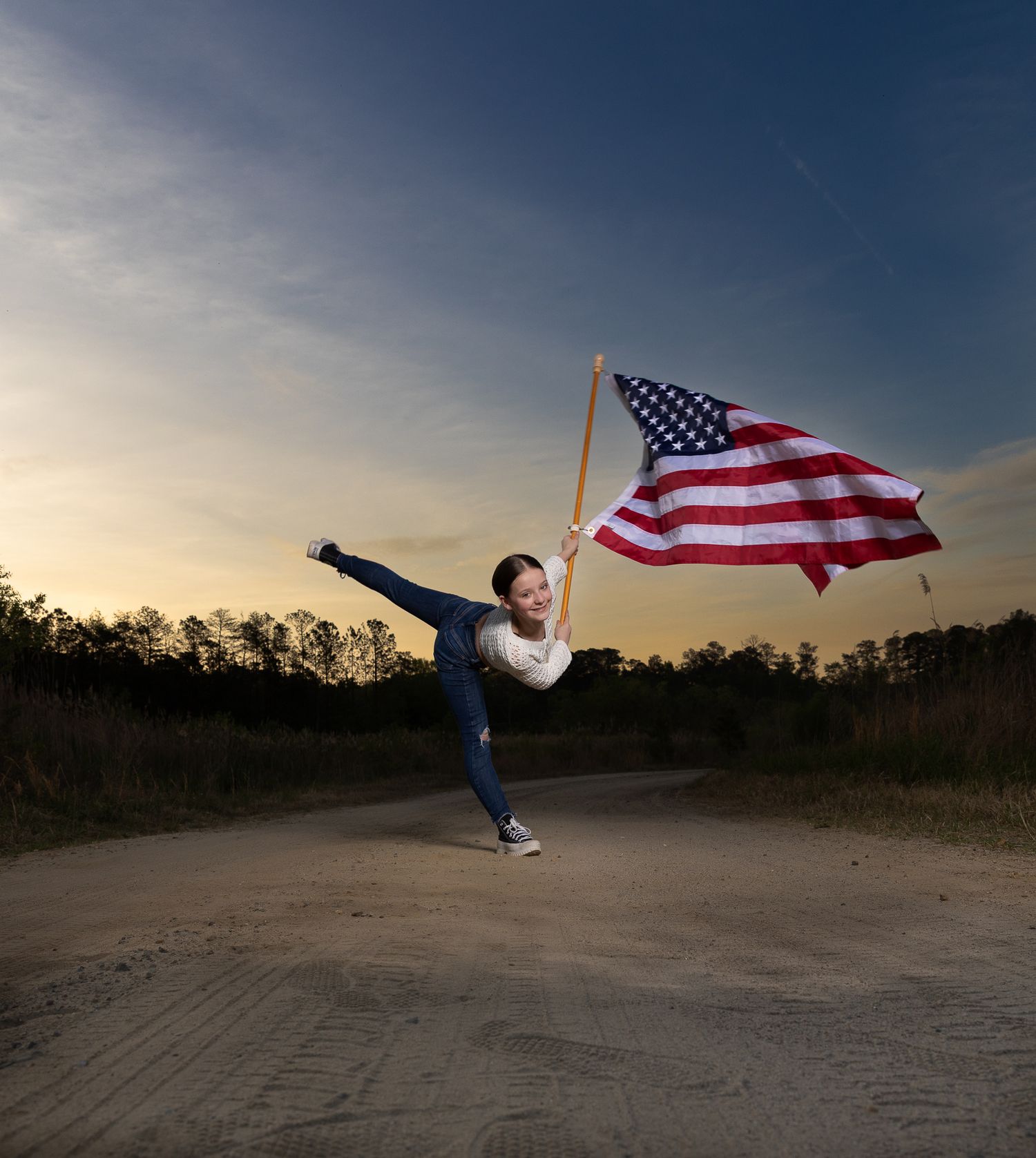 A joyful woman leaps in a golden field holding an American flag, wearing a floral outfit under a blue sky.