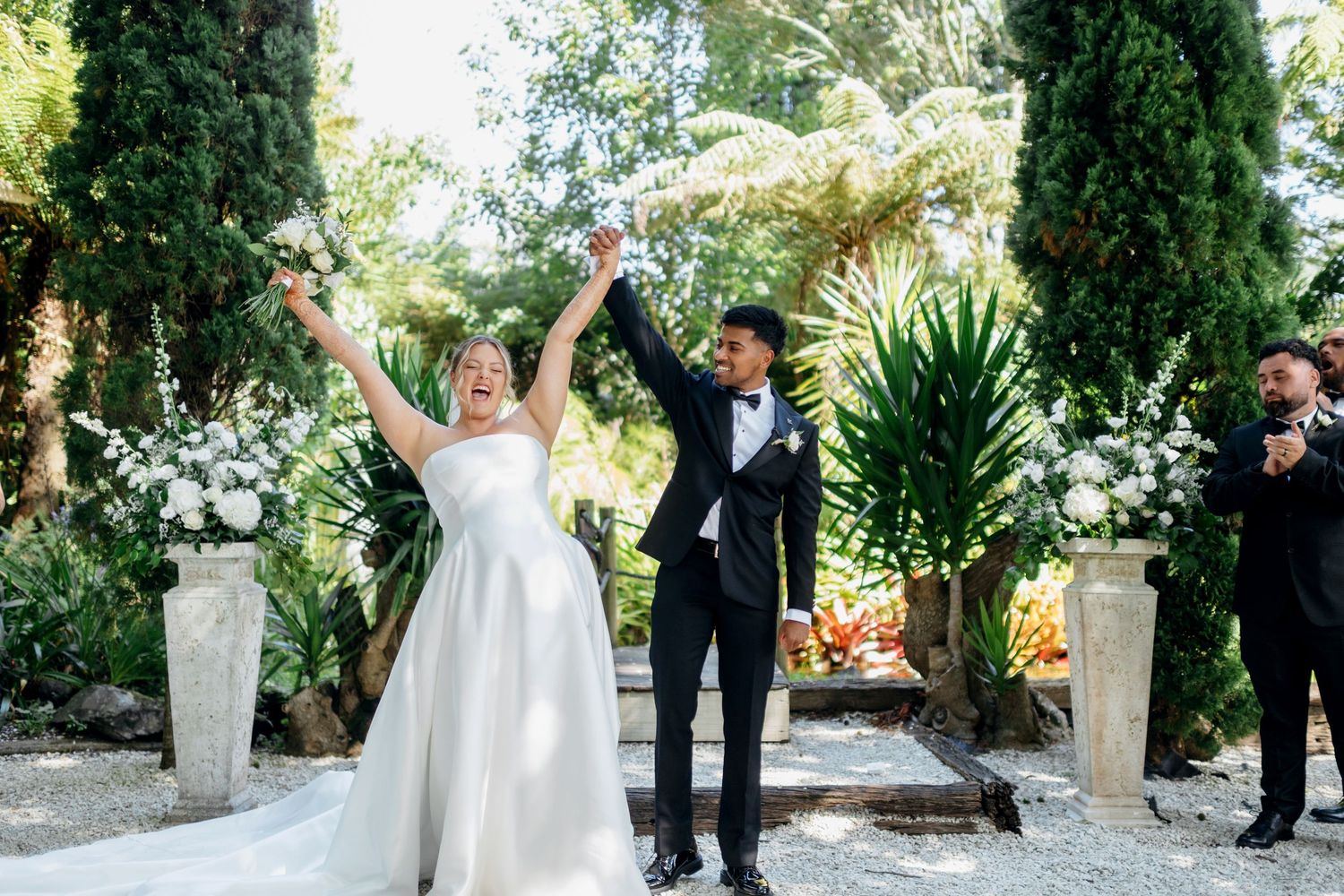 Joyful bride and groom raising hands at outdoor garden wedding ceremony surrounded by lush greenery and white floral arrangements.
