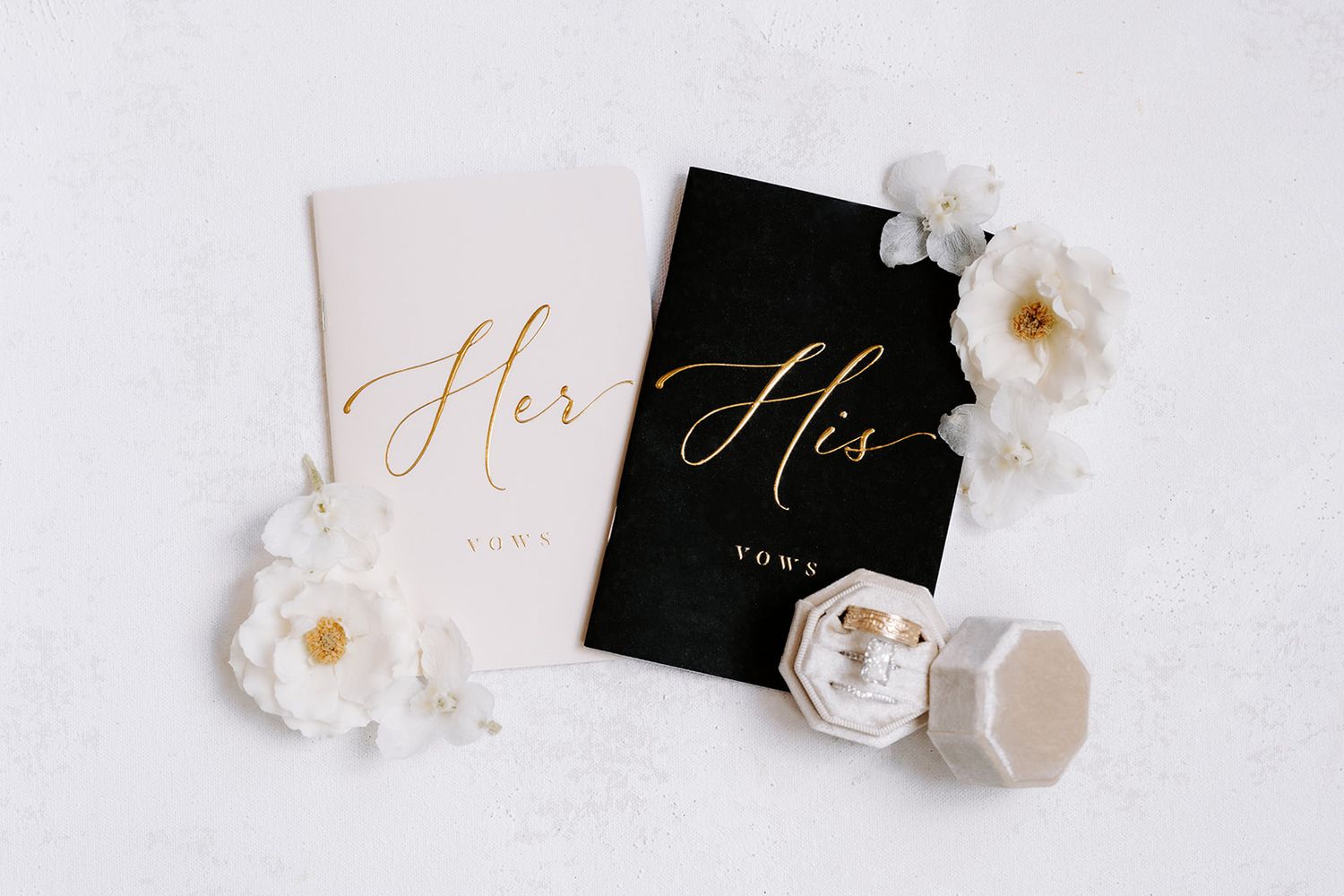 His and Her wedding vow books in black and white with gold script, surrounded by white flowers and a ring box.