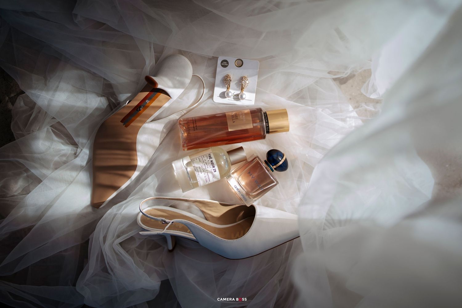 Bridal flat lay with white heels, perfume bottles, earrings, and accessories arranged on a flowing white veil.