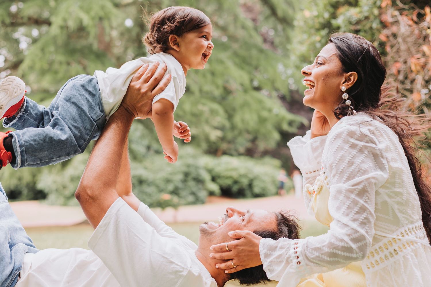 A joyful family outdoors, with a man lifting a laughing baby while a smiling woman in a white lace dress looks on.