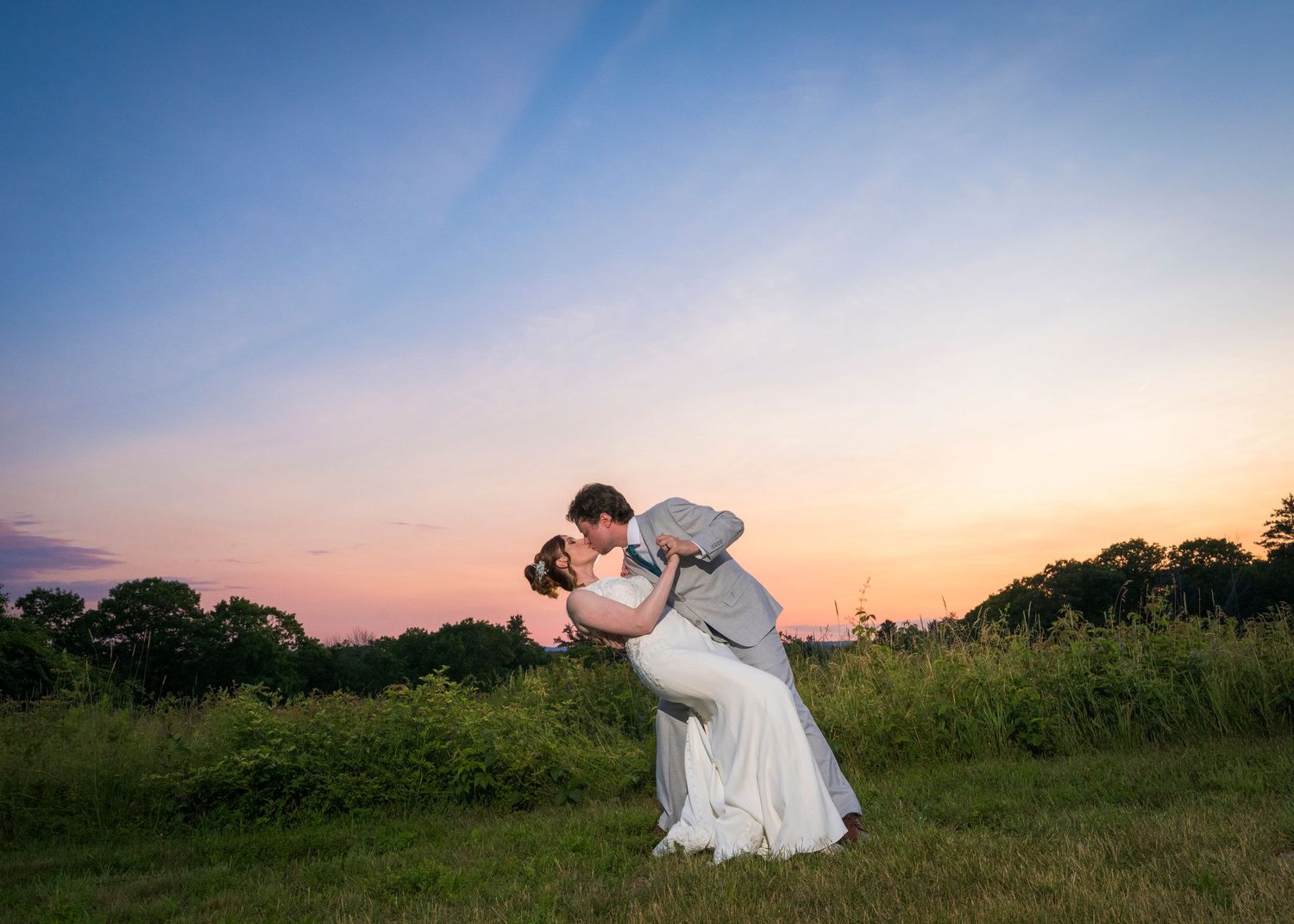 Bride and groom stand on stone steps holding hands before a white farmhouse with green shutters on their wedding day.