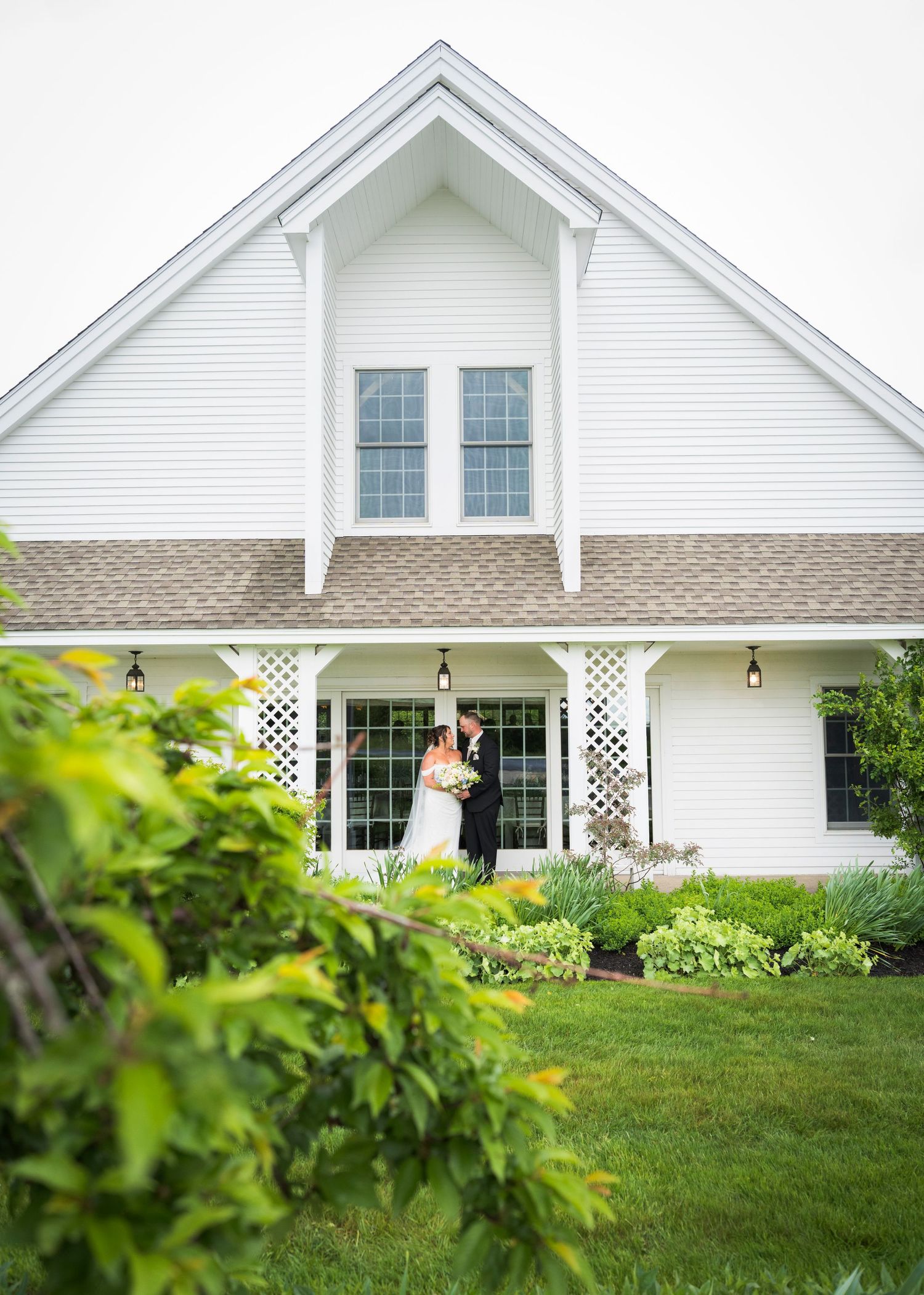 Bride and groom share a romantic kiss on the porch of a charming white farmhouse venue surrounded by lush greenery.