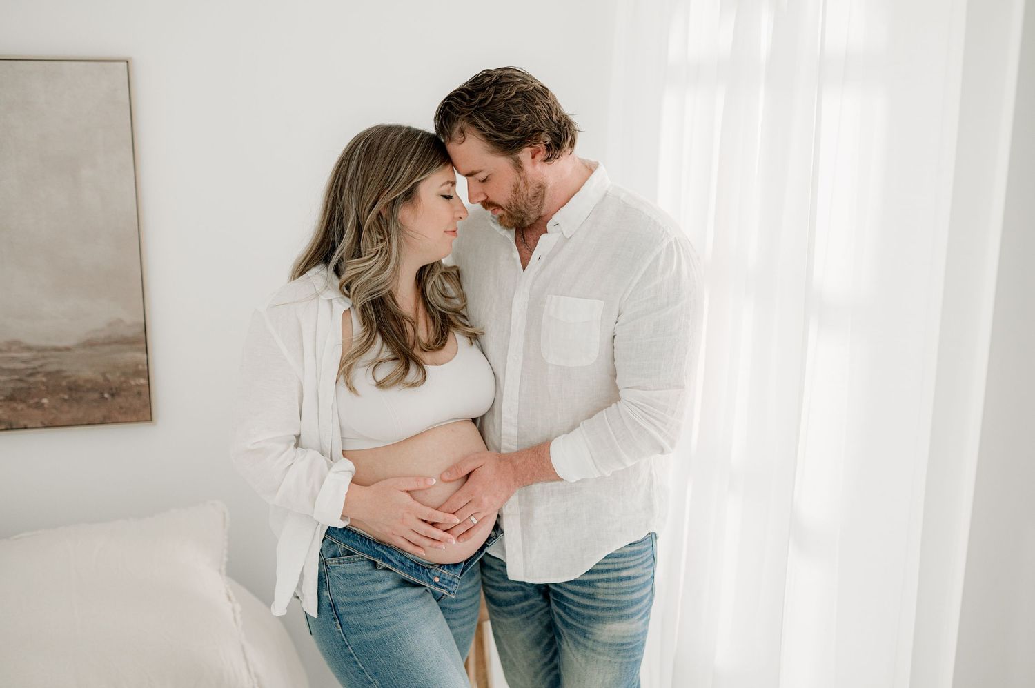 A romantic couple in casual white and denim outfits embrace intimately against a light neutral background.