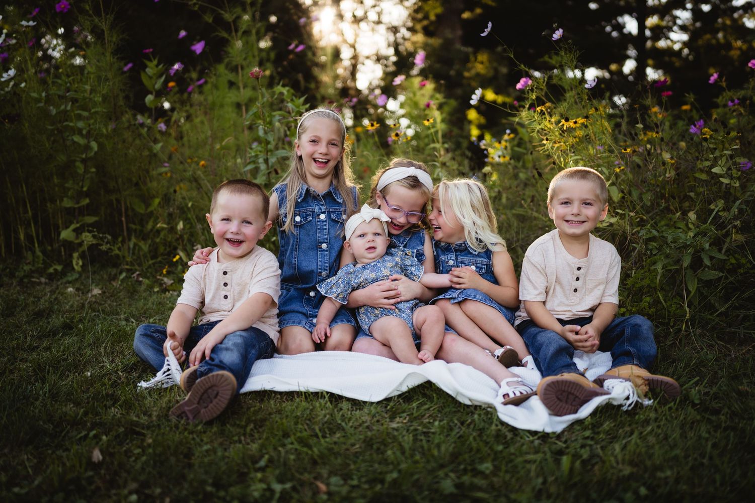 Six young children sitting and smiling on a low tree branch in a lush green park during golden hour.