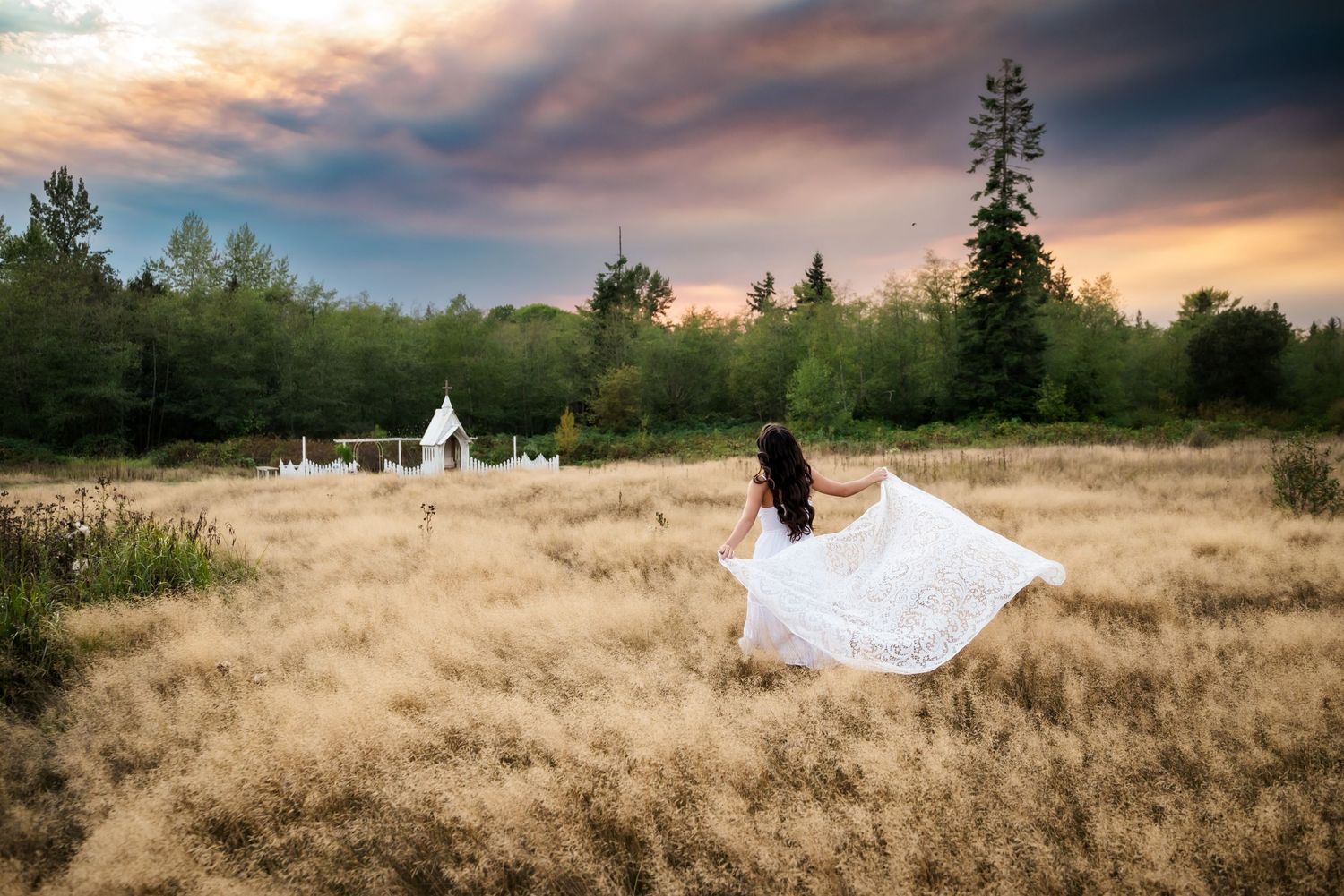 Senior girl wearing white dress in golden field, backed by a rainbow sky, with lace flowing behind her