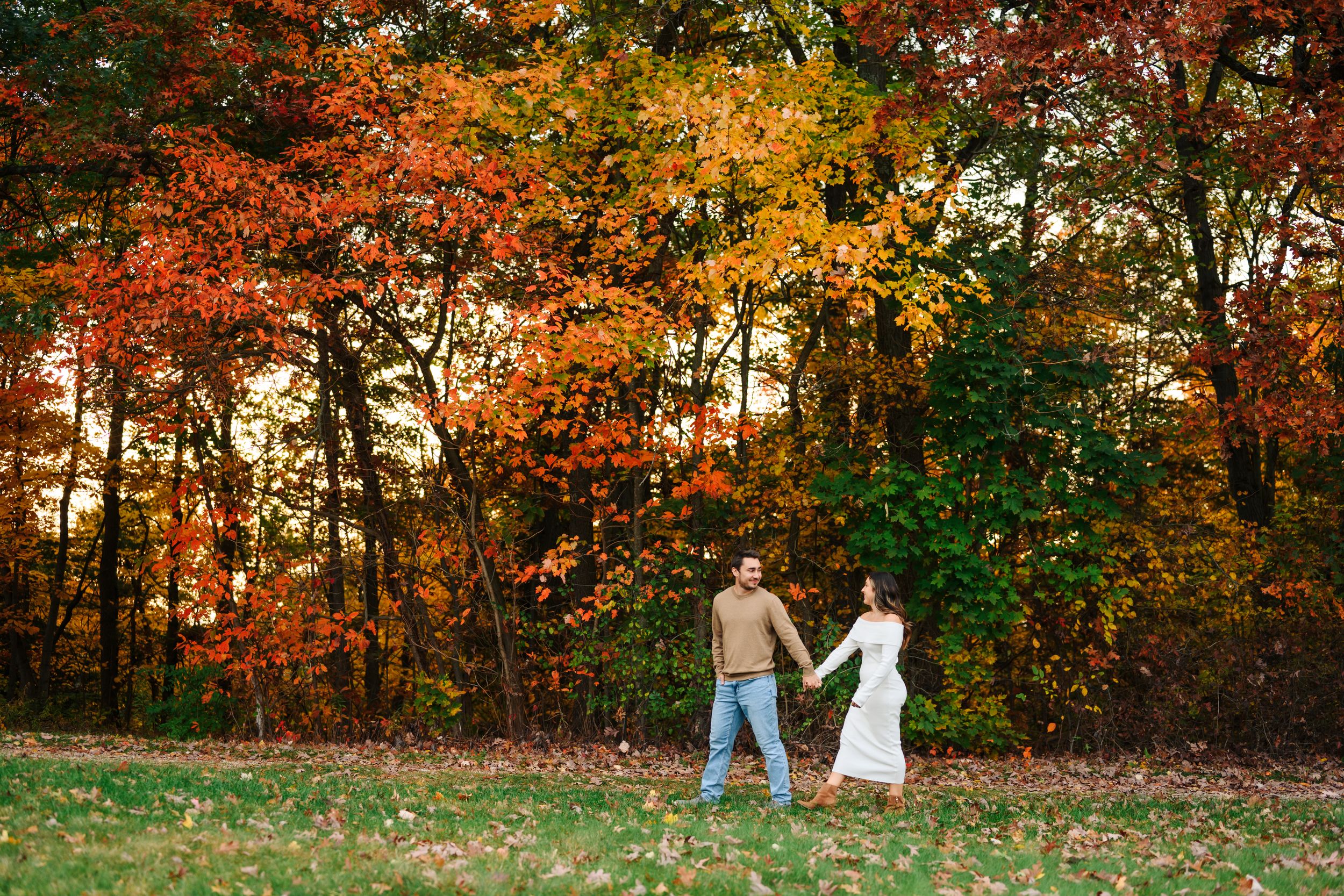 A couple walks hand in hand through vibrant autumn foliage with red and yellow trees in the background.