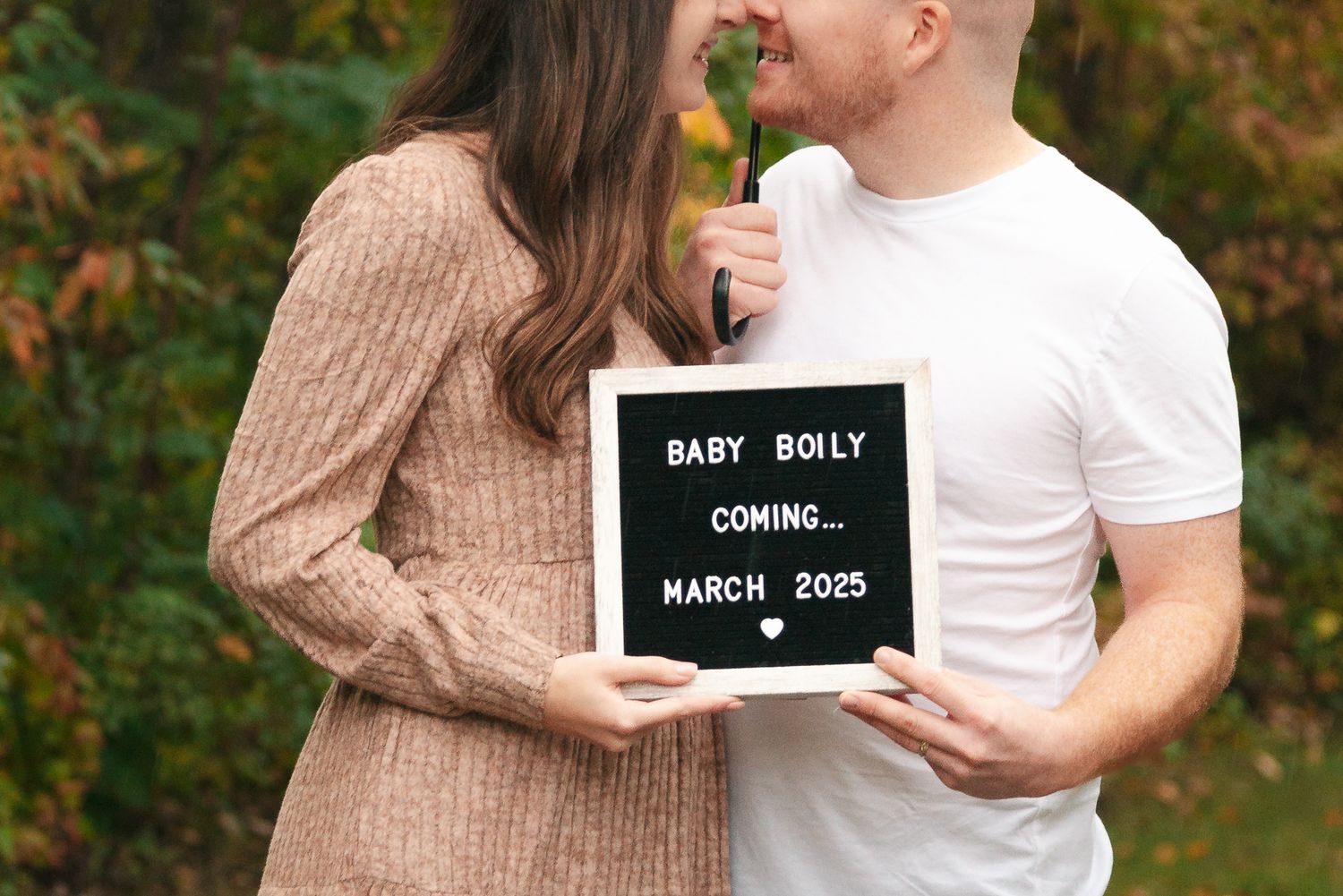 A couple holding a letter board announcing Baby Rolly coming in March 2023 against autumn foliage.
