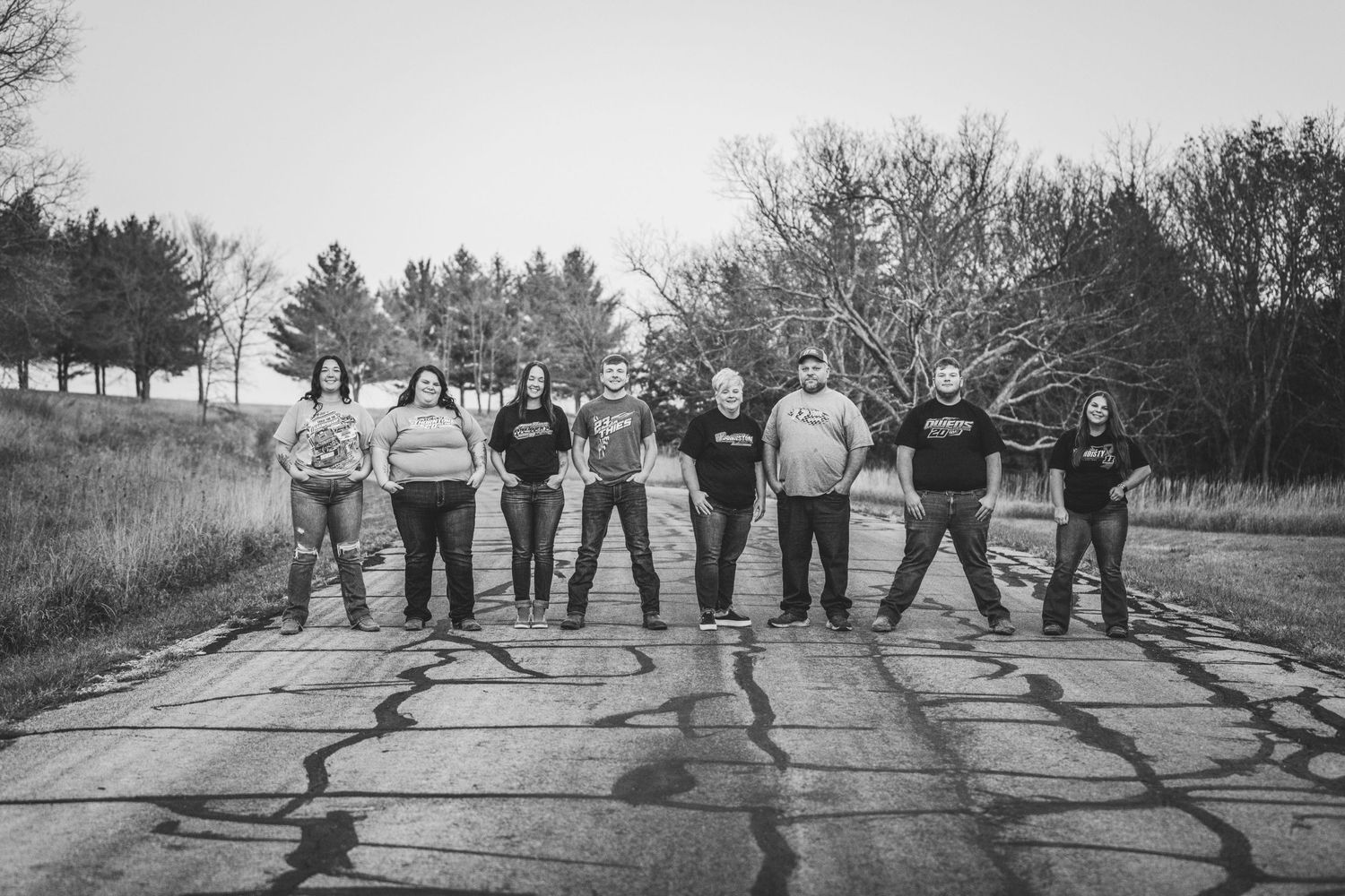 A group of people stand together on a cracked rural road in black and white, with trees lining the pathway in the background.