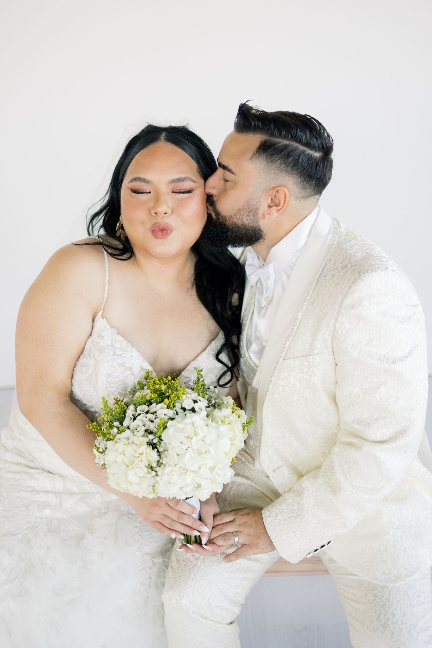 Wedding couple in matching white outfits share sweet moment while holding a bouquet of white flowers and greenery.