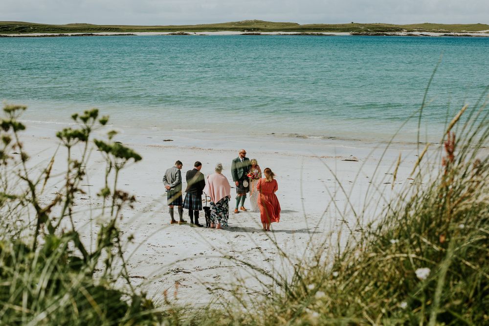 A group stands together on a white sandy beach with turquoise waters and green coastal vegetation in the foreground.