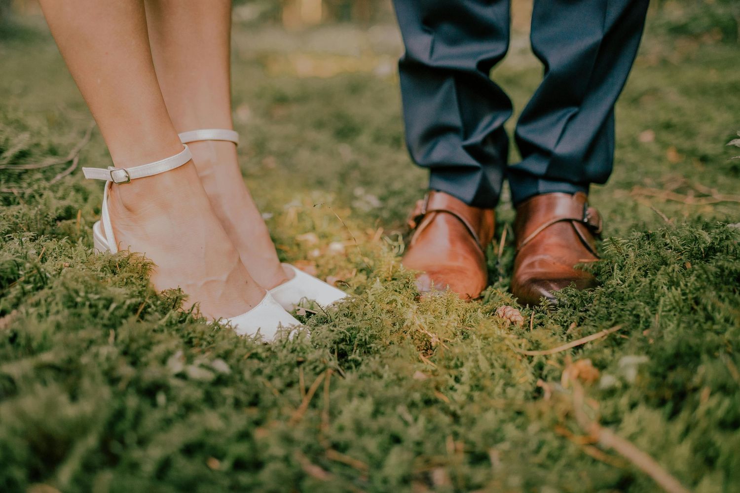 Low angle view of contrasting elegant white bridal shoes and brown leather dress shoes standing in grass.