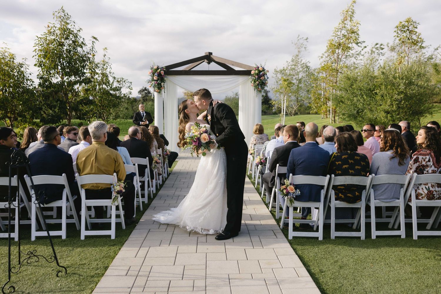 An outdoor wedding ceremony under an arch with guests seated in white chairs on a stone patio surrounded by trees.