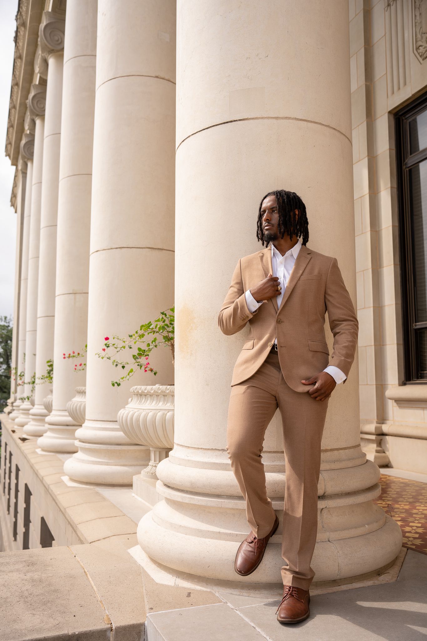Stylishly dressed individual in beige suit poses against classical stone columns of an elegant building.