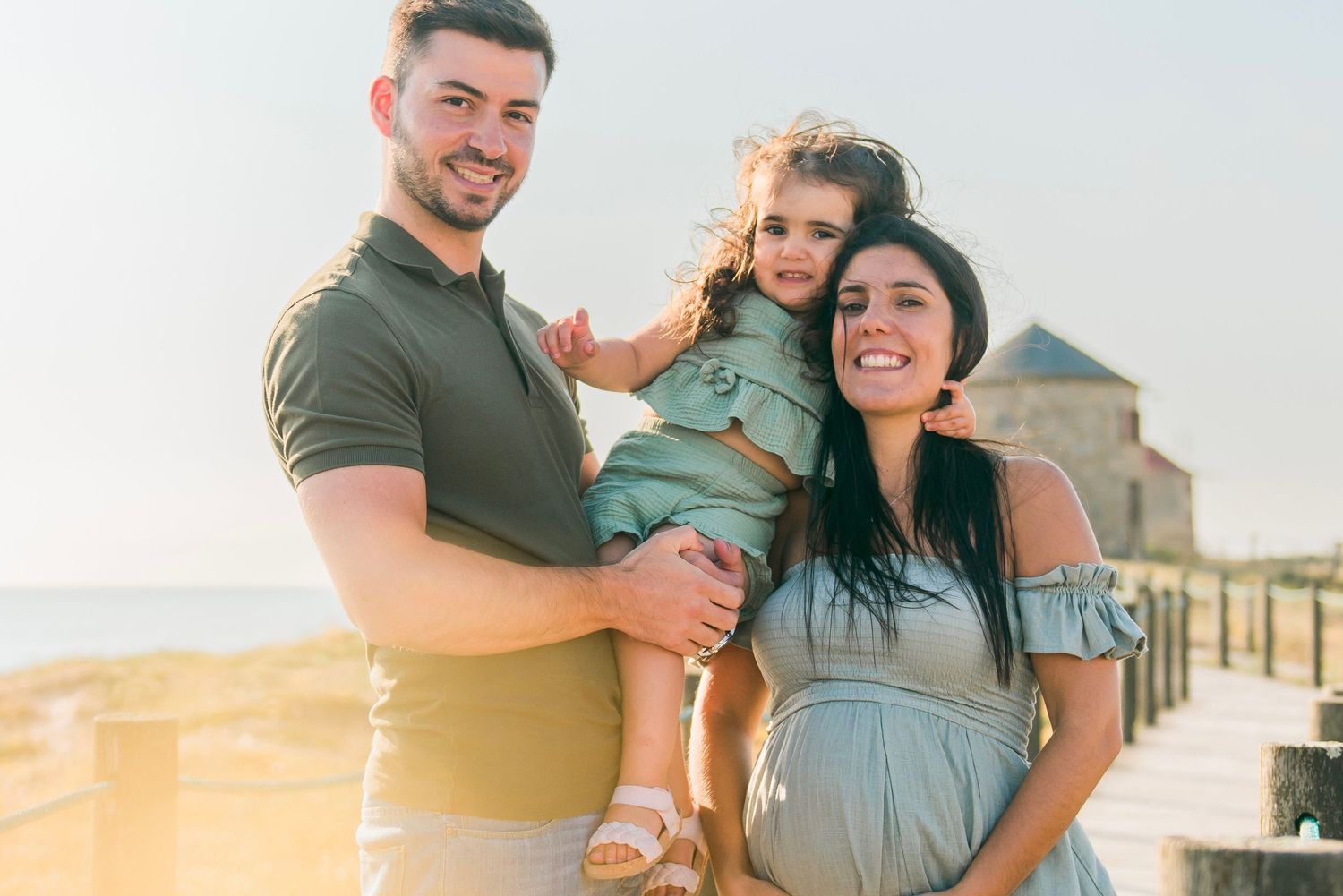 Happy family with pregnant mother, father, and toddler posing at golden hour by sea.