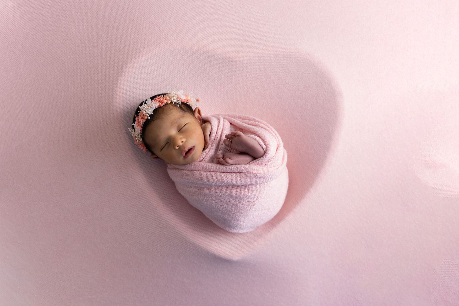 Newborn baby wrapped in soft pink swaddle cloth sleeping peacefully on pink background with floral headband.