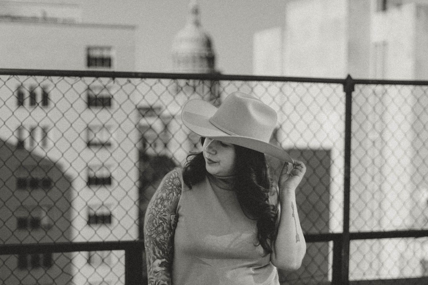 Woman in sleeveless top and sun hat poses on urban rooftop with chain link fence and buildings in black and white photo.