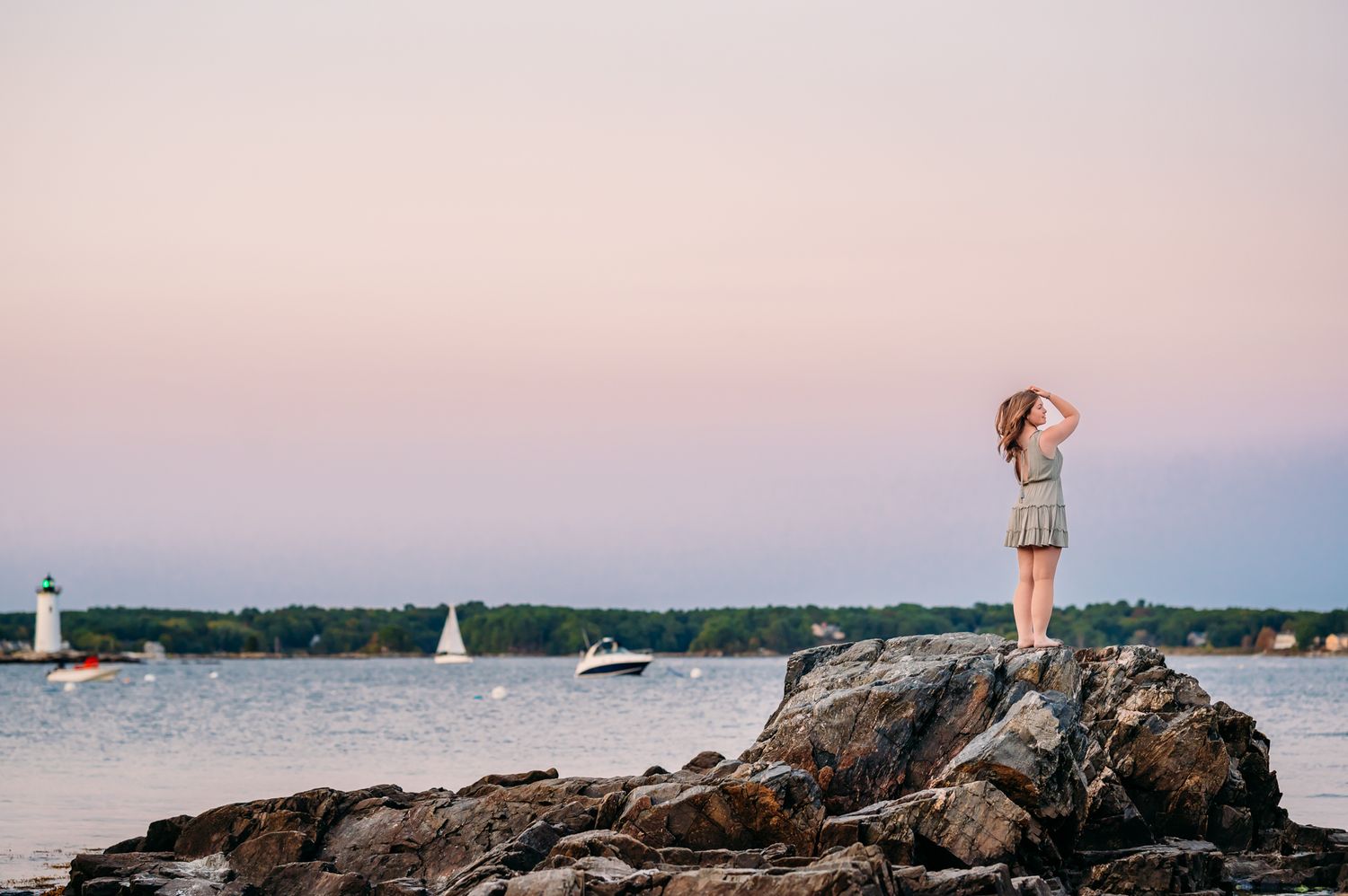 Someone stands on rocky coastal outcrop overlooking harbor with sailboats during sunset at Maine coastline.
