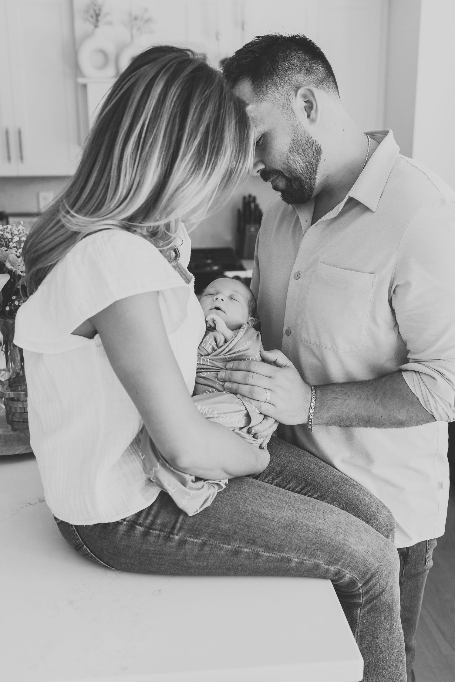 black and white image of a colts neck couple embracing their new baby girl while standing their kitchen.