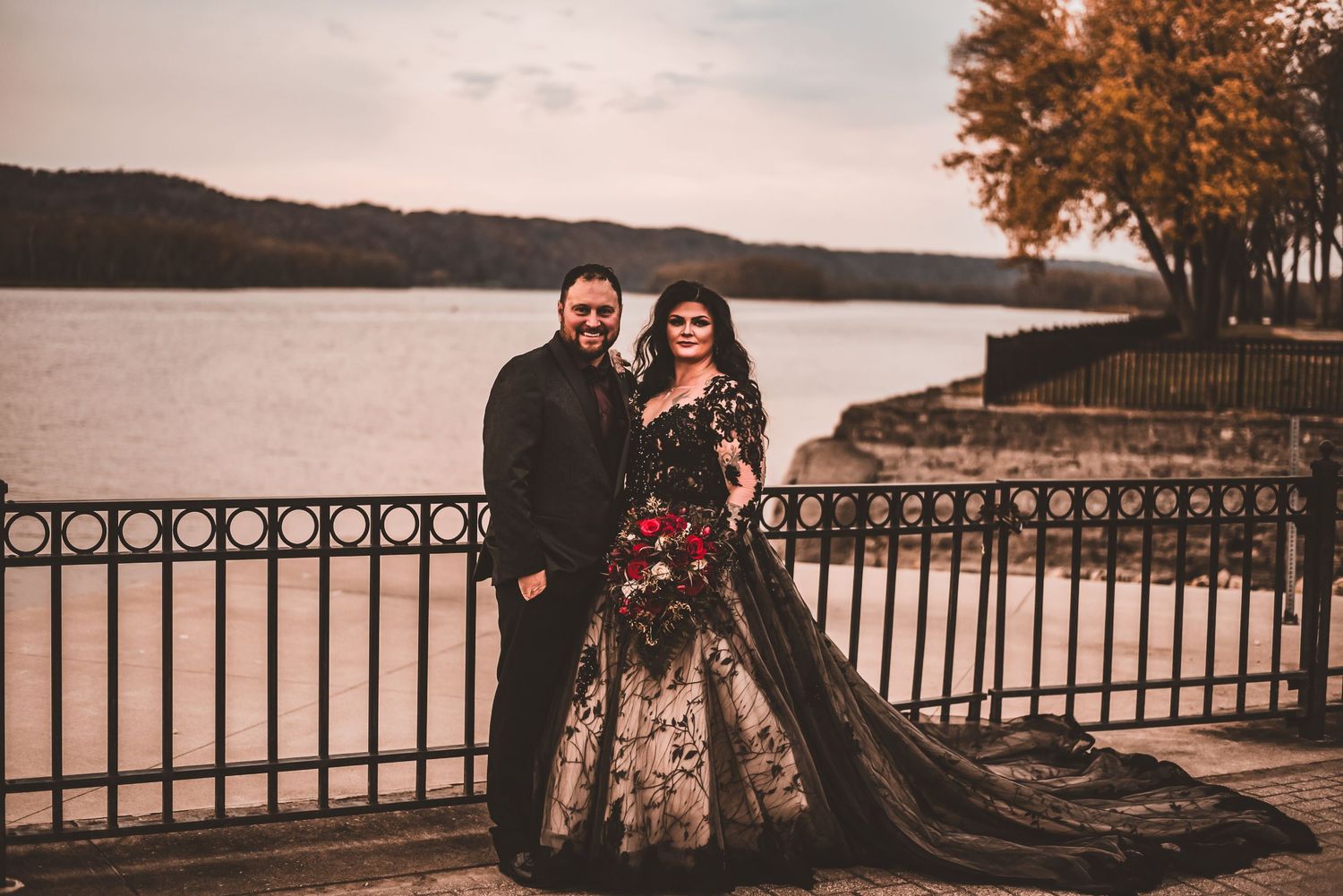 A couple poses by a lakeside railing during autumn with golden trees in the background.