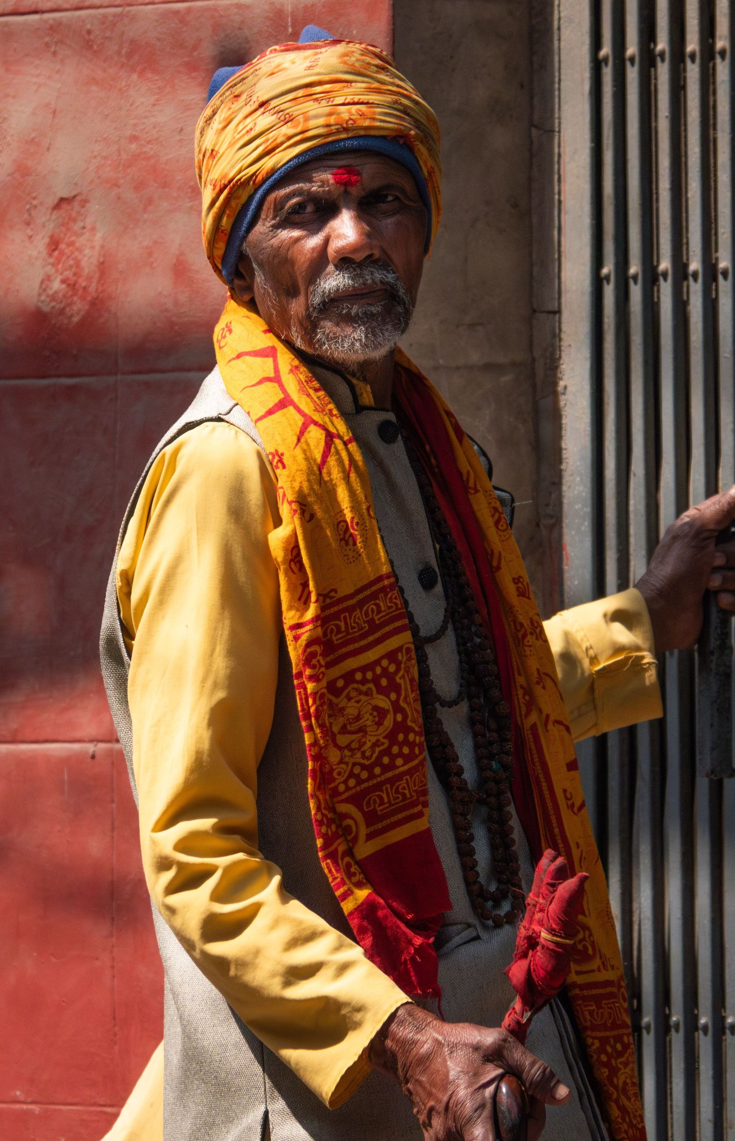 A person in traditional orange and yellow attire with turban stands against a red wall.
