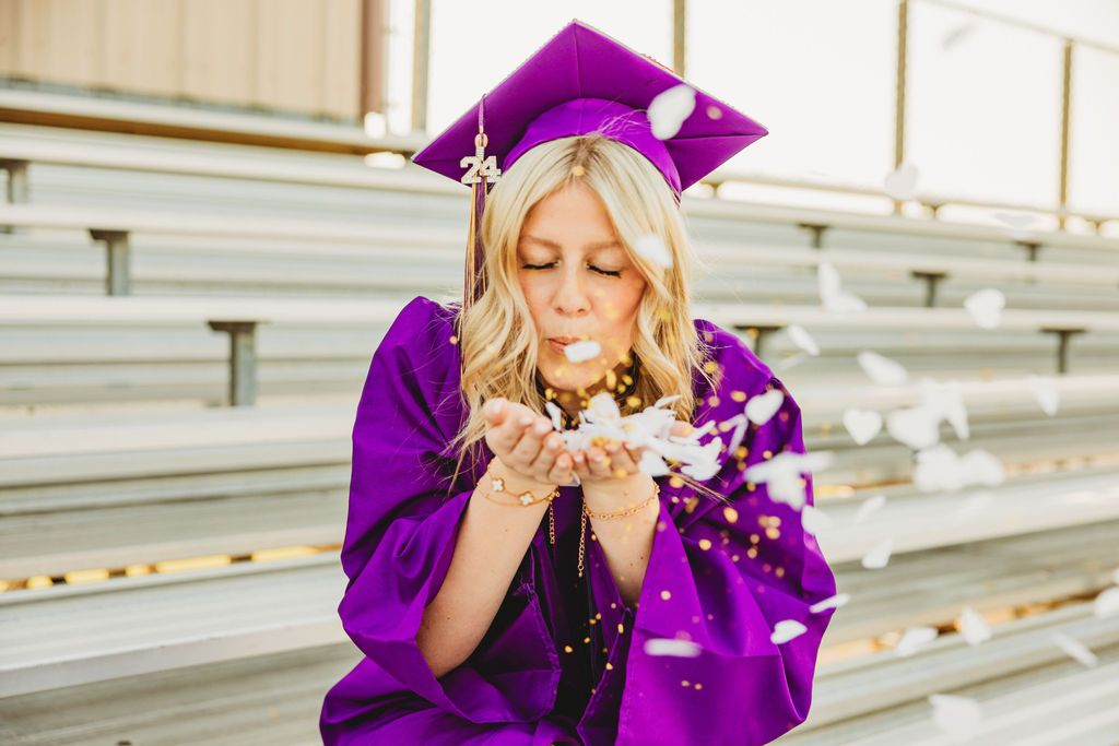 Graduate in purple gown celebrates with glitter confetti on outdoor bleachers.
