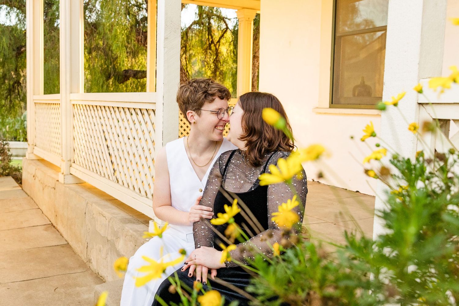 A couple embraces on a porch among blooming yellow flowers and lattice railing on a sunny day.