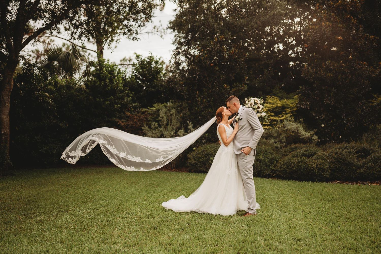 Wedding portrait with flowing white veil against green garden background.