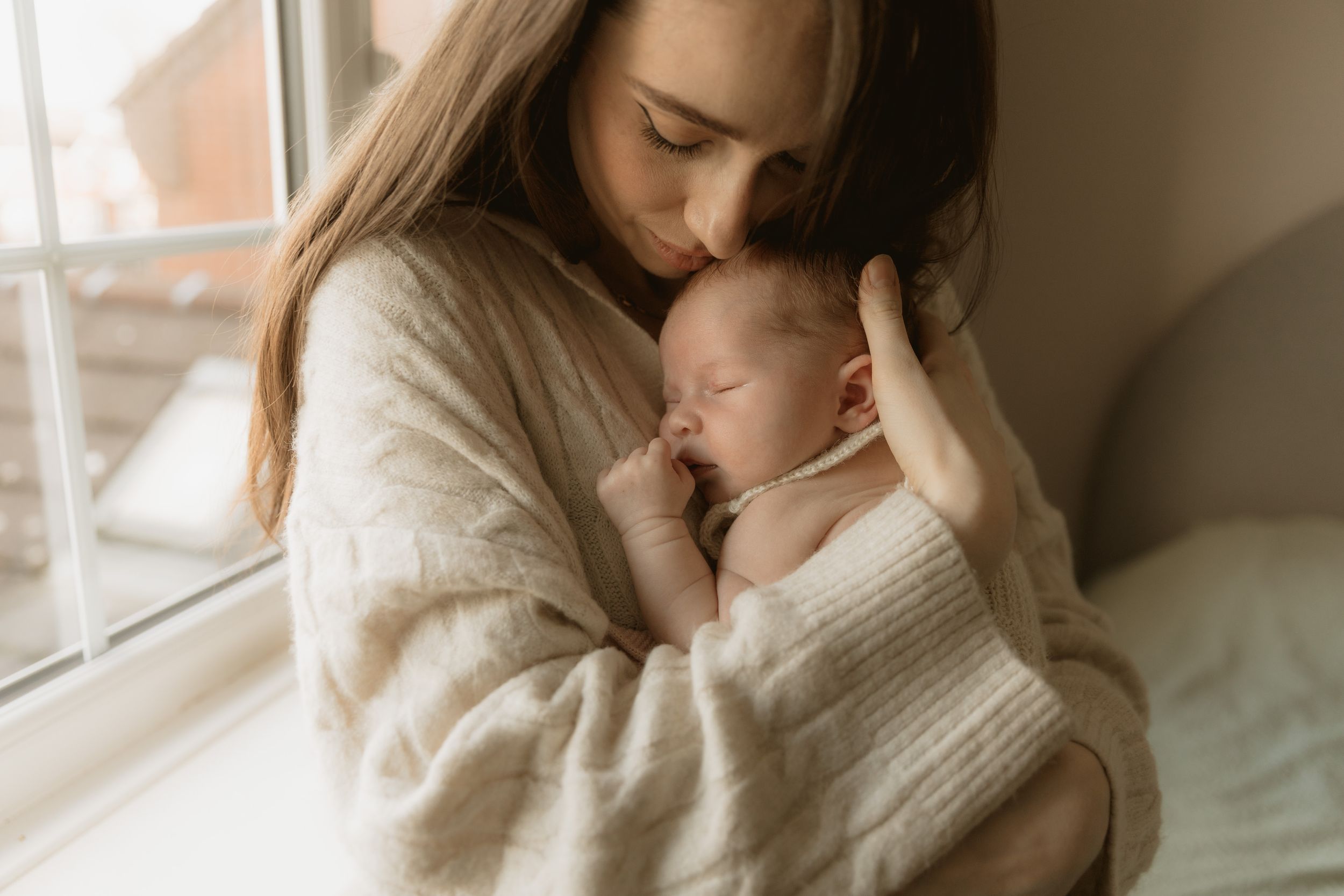 A parent tenderly embraces their small infant in a bright sunlit room near a window with soft white textiles.
